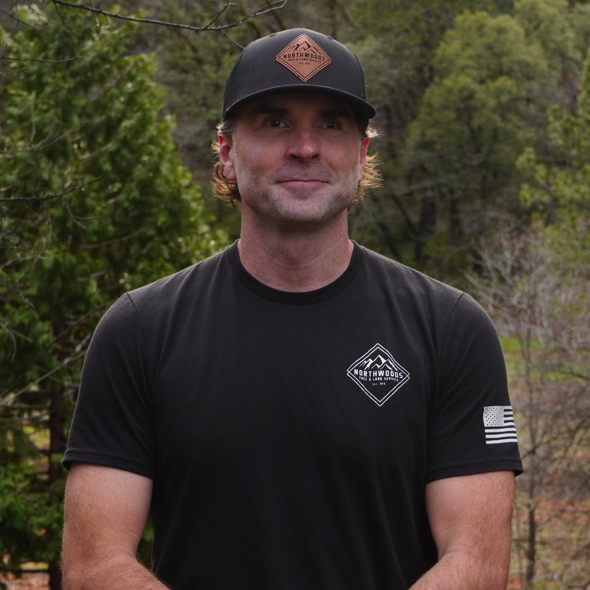 A person in an orange jacket sitting atop a tall, cut tree trunk in a forested area with leafless trees and dry grass, overlooking hilly terrain with winding roads.