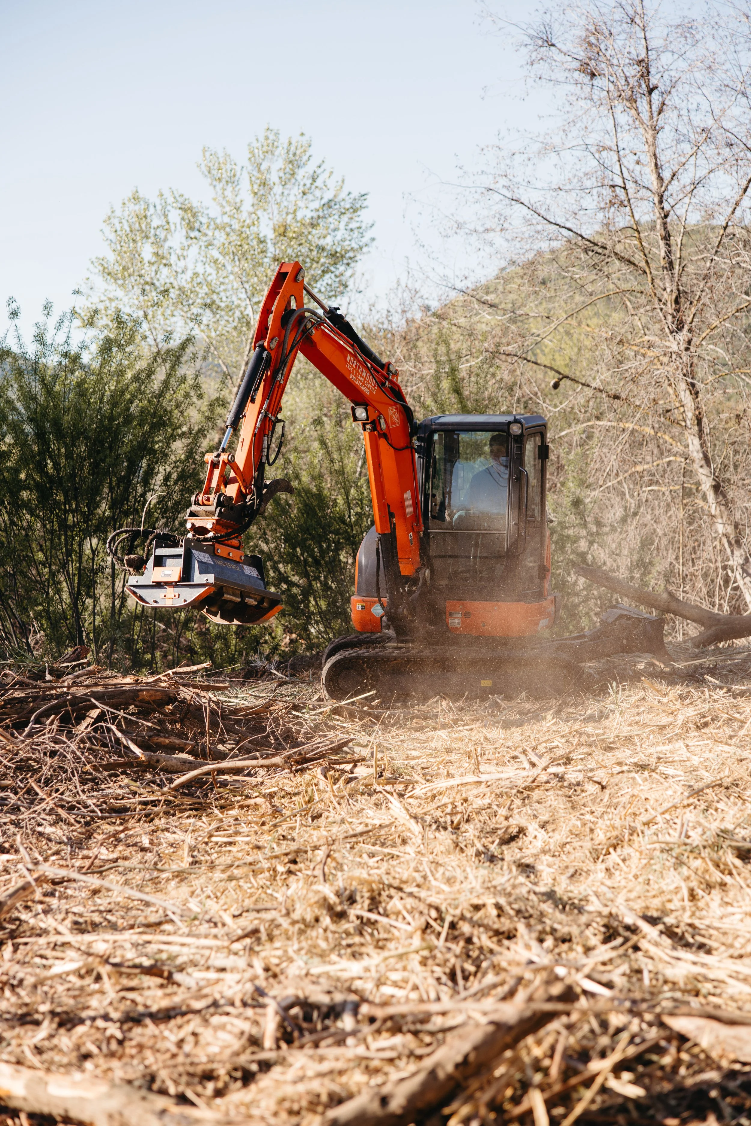 A construction excavator operating on a cleared outdoor area with wood debris and trees in the background.