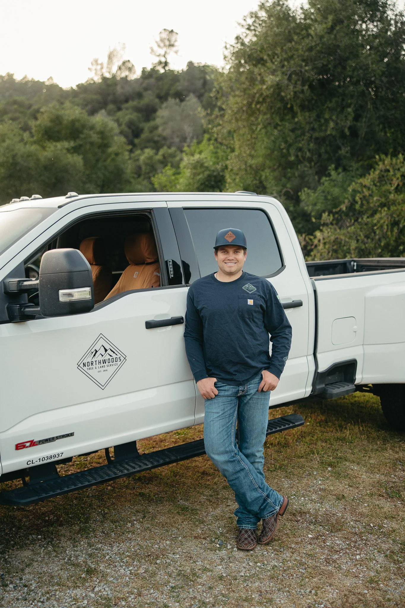 A man in a black long-sleeve shirt and jeans standing beside a white pickup truck with a Northwoods Tree & Land Service logo, outdoors with trees and hills in the background.