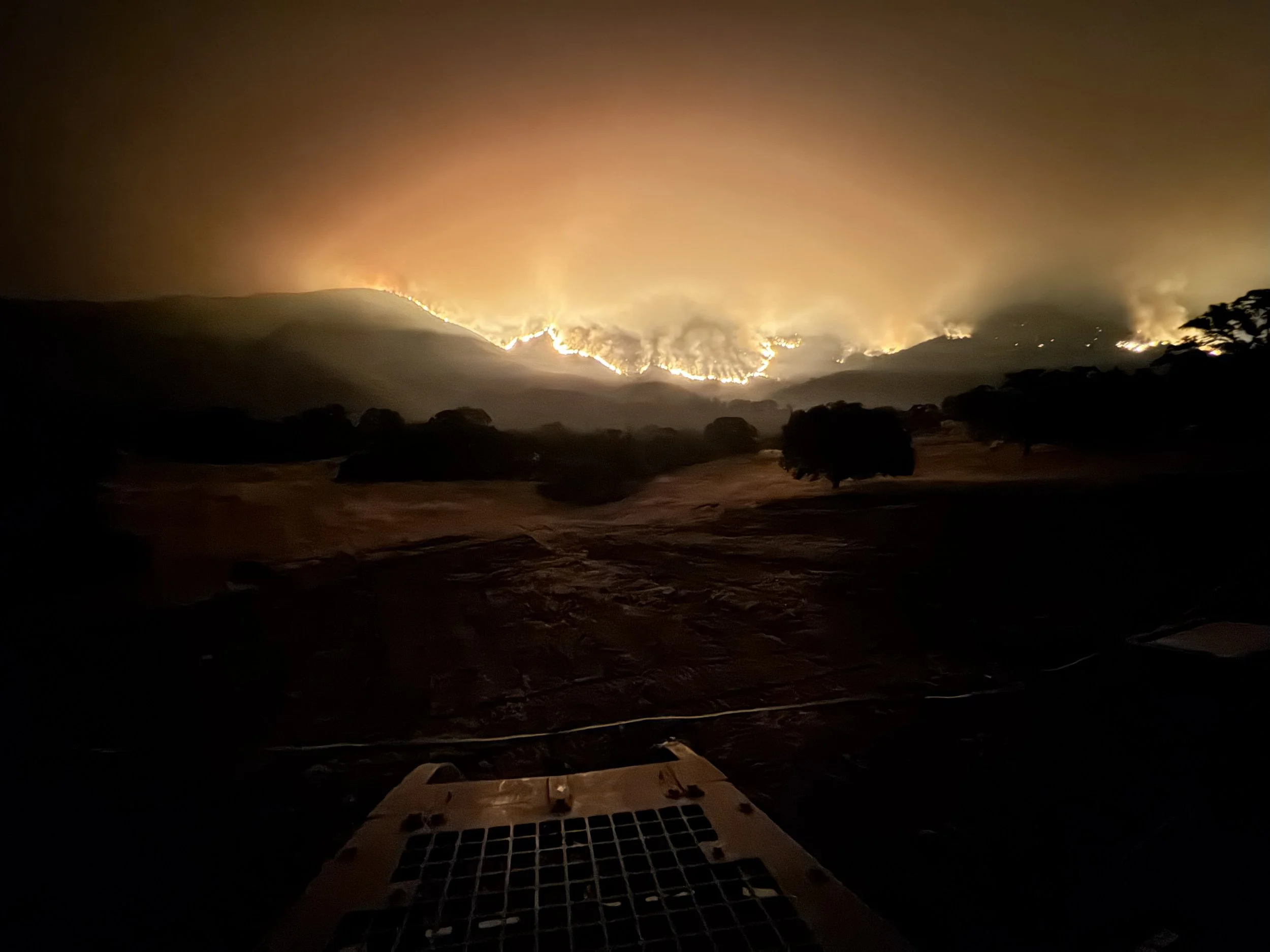 Nighttime view of a wildfire on a mountain, with bright flames and smoke illuminating the sky, seen from a dark area with trees and a drone or camera equipment in the foreground.