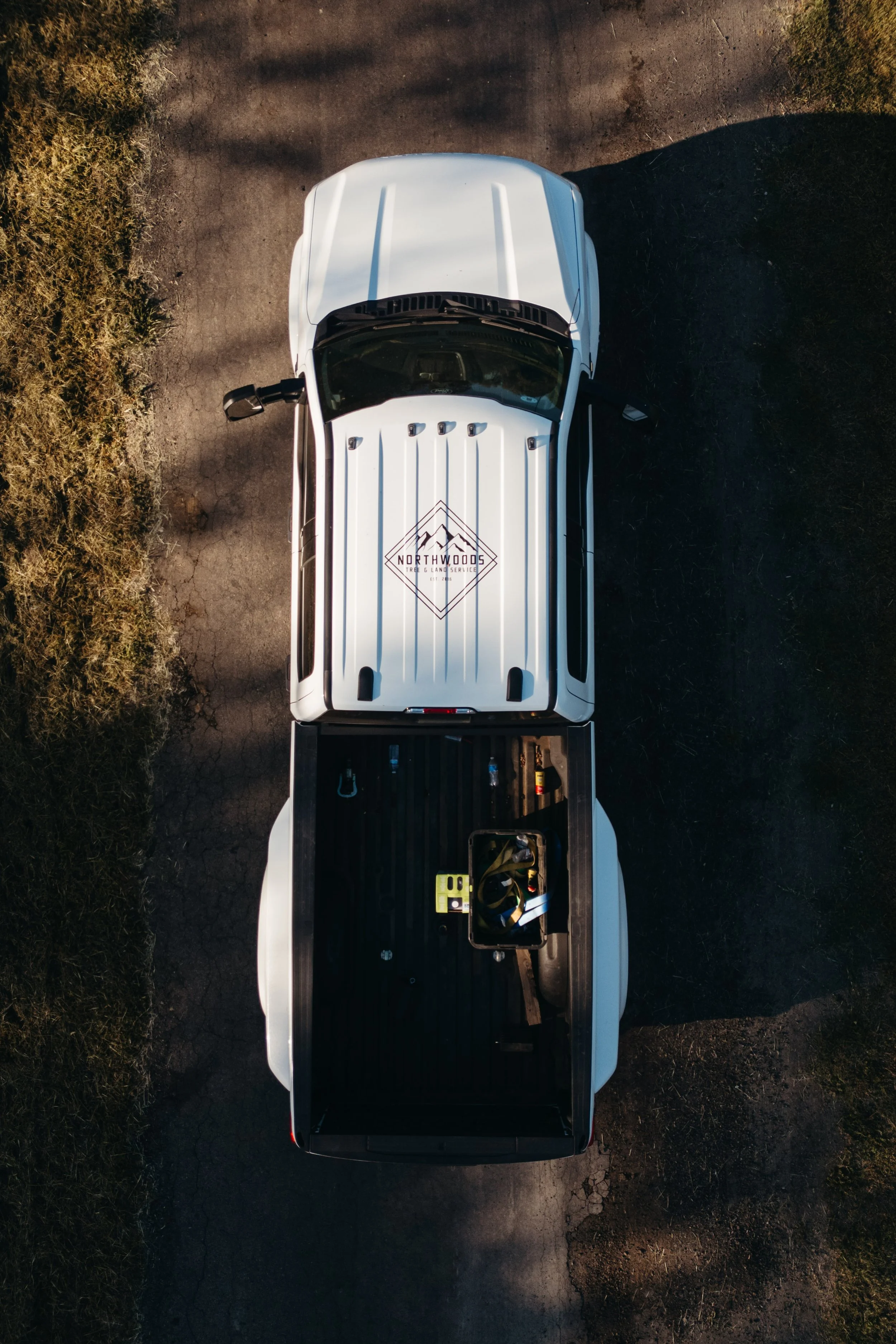 Aerial view of a white pickup truck with an open bed on a rough, cracked road, surrounded by grass and trees. The truck's roof has a logo for 'Northwoods Tree and Land Service'.
