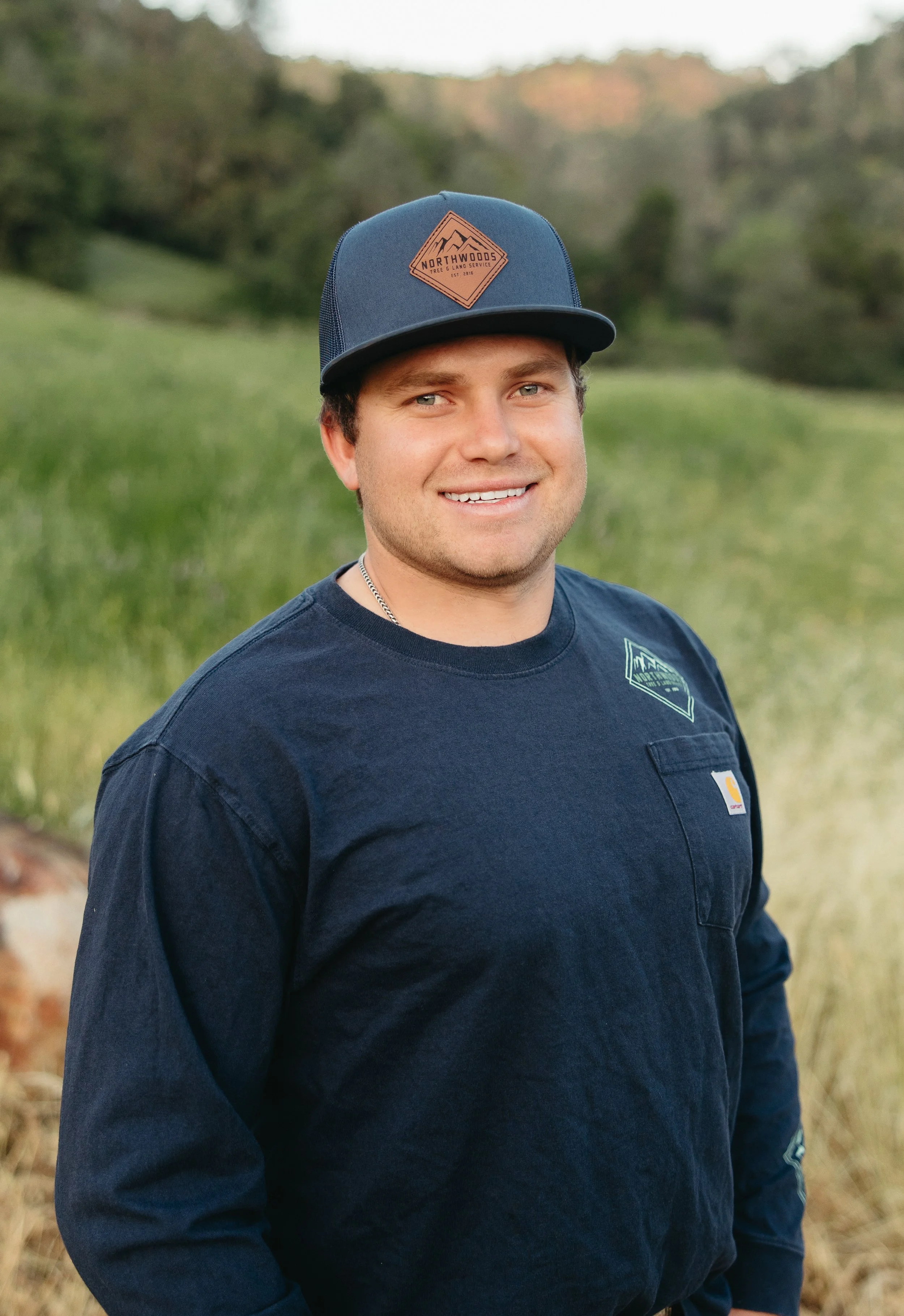A young man with dark hair smiling outdoors, wearing a navy blue cap and matching long sleeve shirt, standing in a green field with trees in the background.