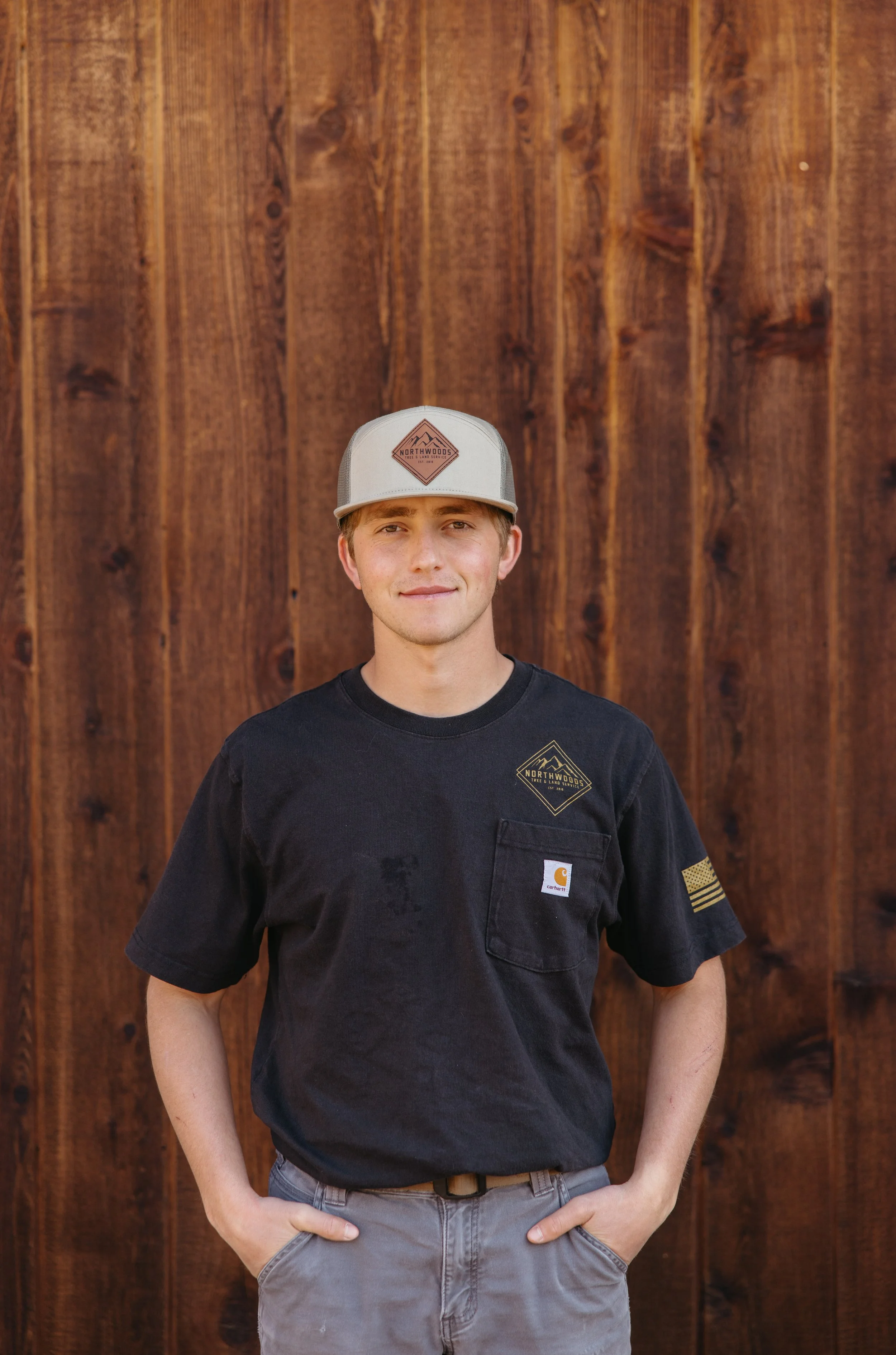 A young man standing in front of a wooden fence, wearing a black Carhartt t-shirt and a gray baseball cap with a Northwoods logo, smiling with his hands in his pockets.