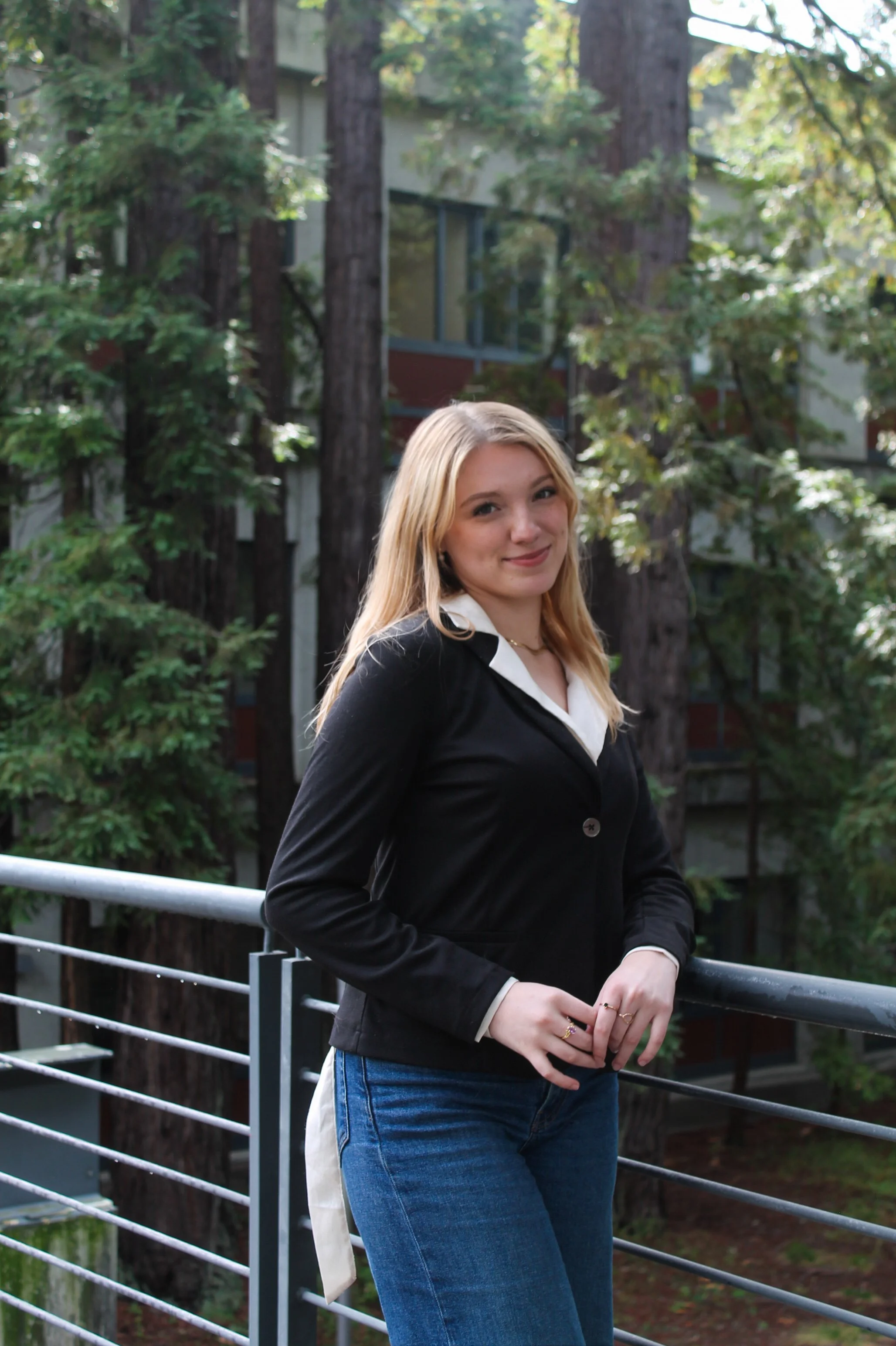 A young woman with blonde hair wearing a black jacket and blue jeans standing on a balcony with trees and a building in the background.