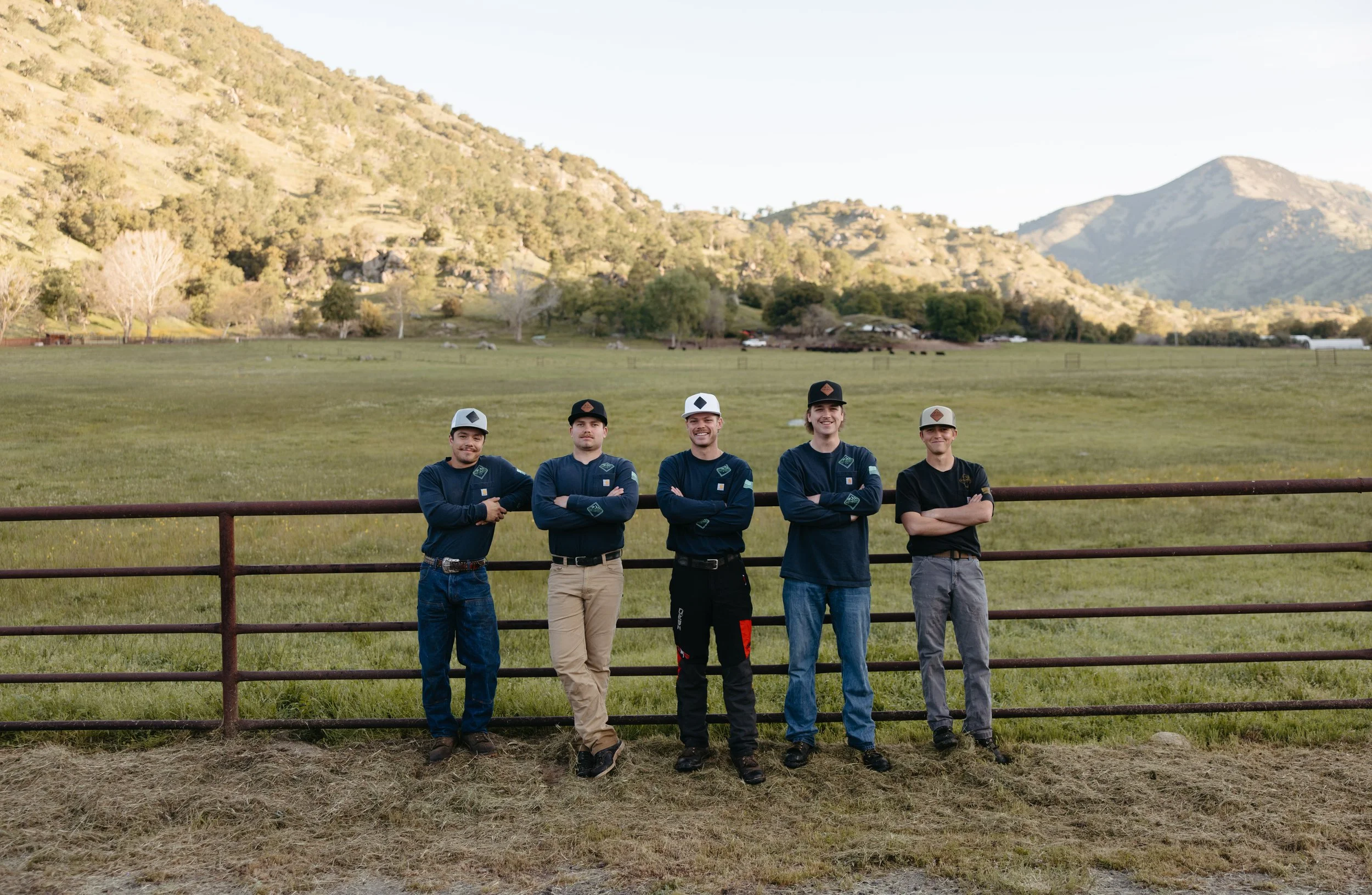 Five young men standing outdoors behind a metal fence, with mountains and green fields in the background, posing with crossed arms.