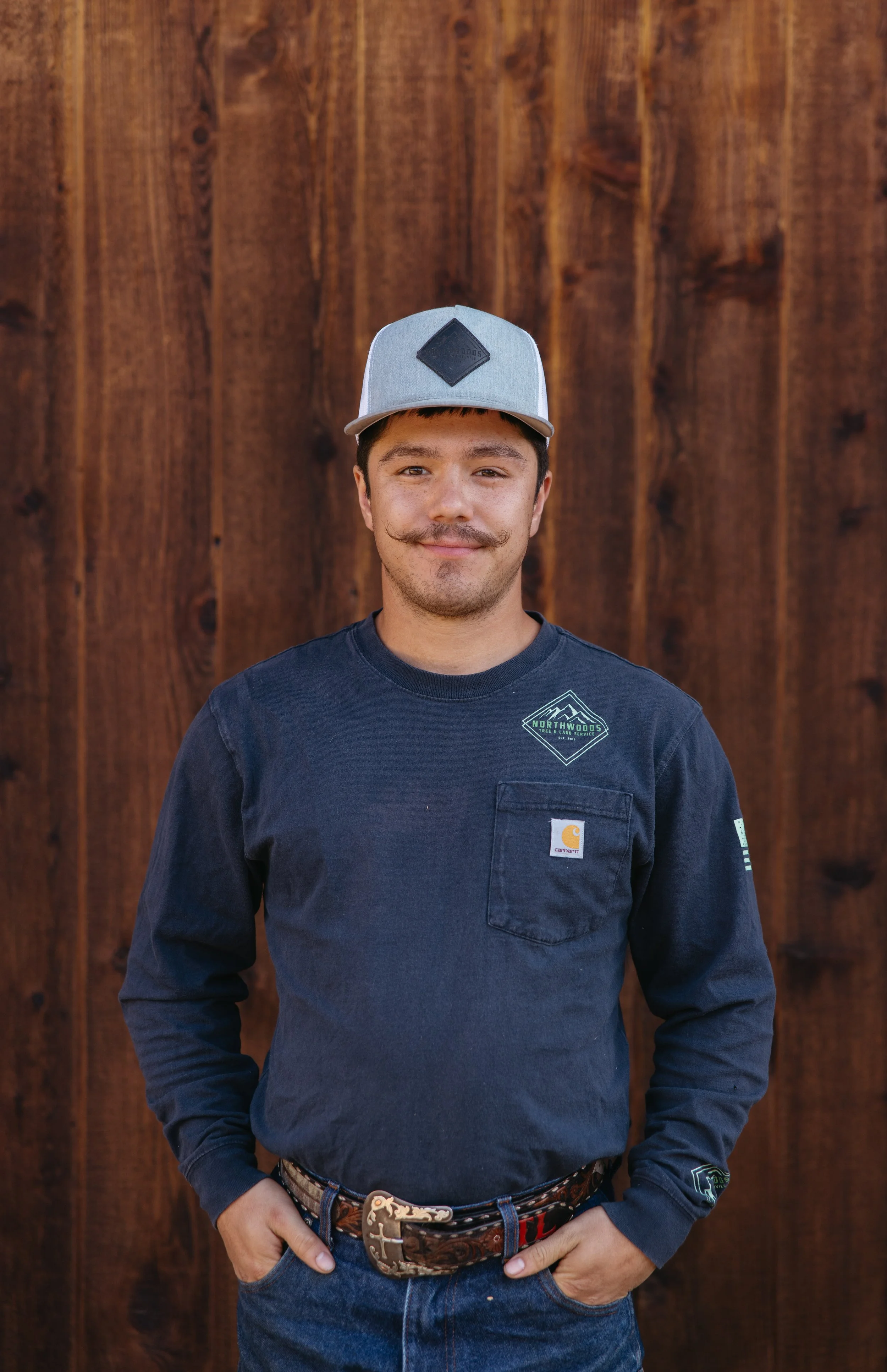 Young man standing outdoors in front of a wooden wall, wearing a light grey hat, a navy blue Carhartt shirt with patches, and a decorative belt.