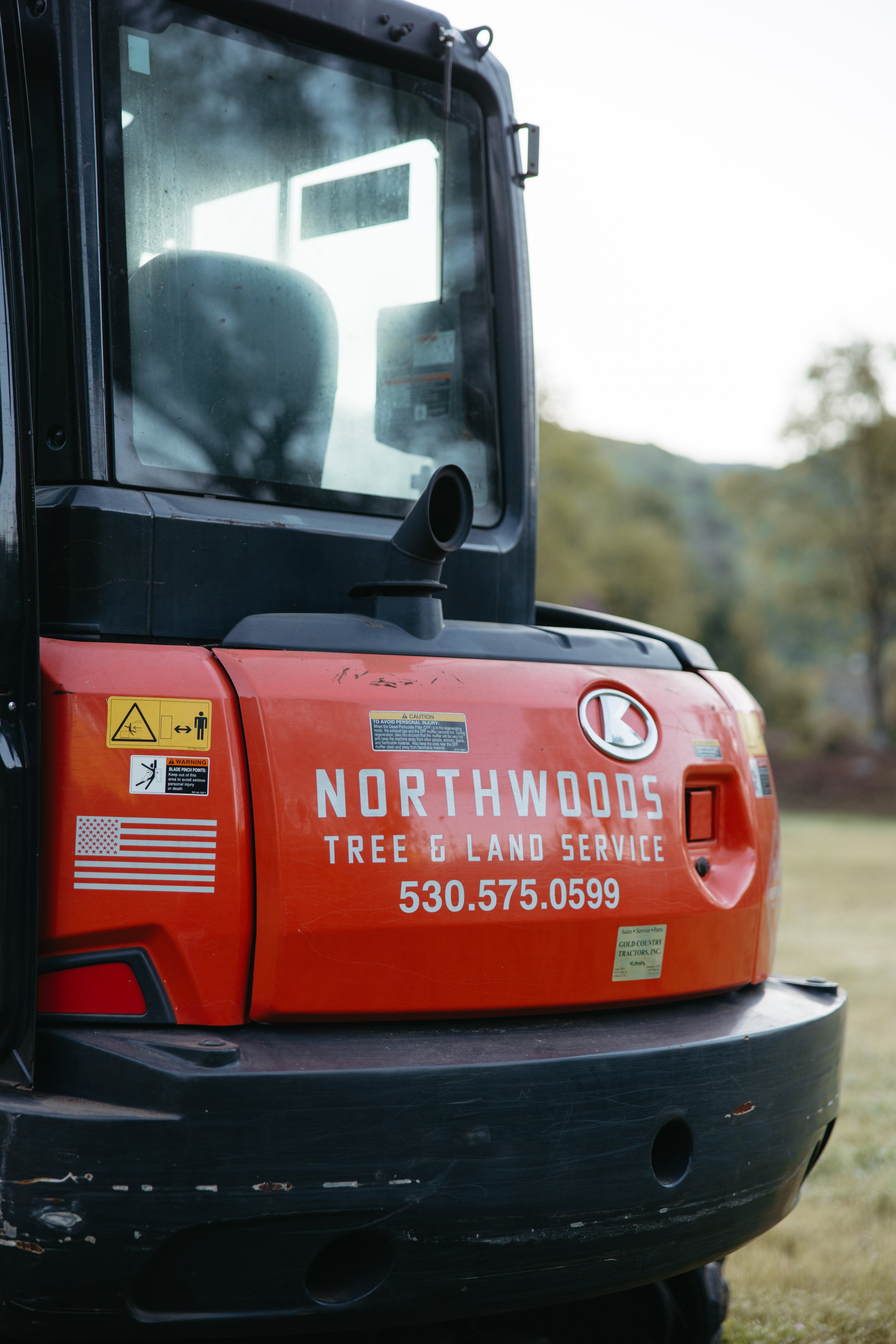 Close-up of a red construction vehicle with Northwoods Tree & Land Service logo, contact number, and safety warning stickers on the side.