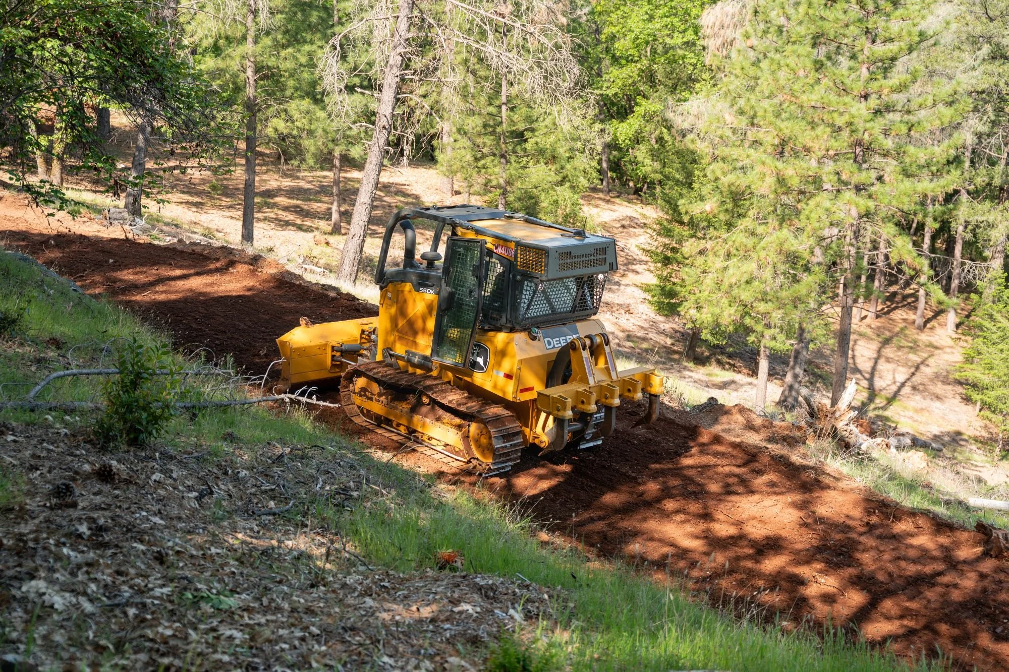 A yellow bulldozer working on a slope in a forested area with green trees.