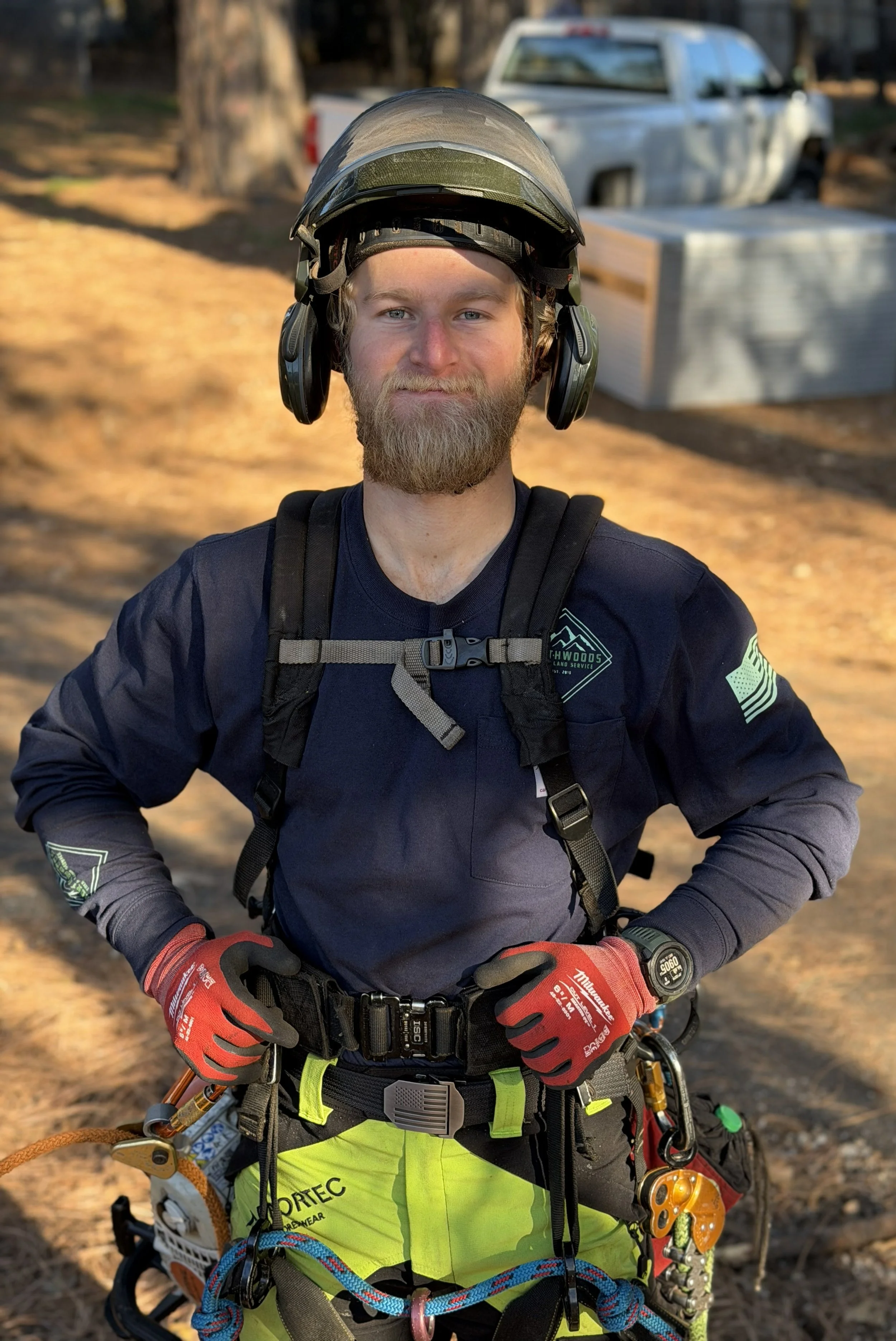 A man with a beard and mustache wearing a helmet with ear protection, red gloves, a black harness, a digital watch, and neon yellow work pants with safety equipment, standing outdoors with a tree, a white truck, and a large silver structure in the background.