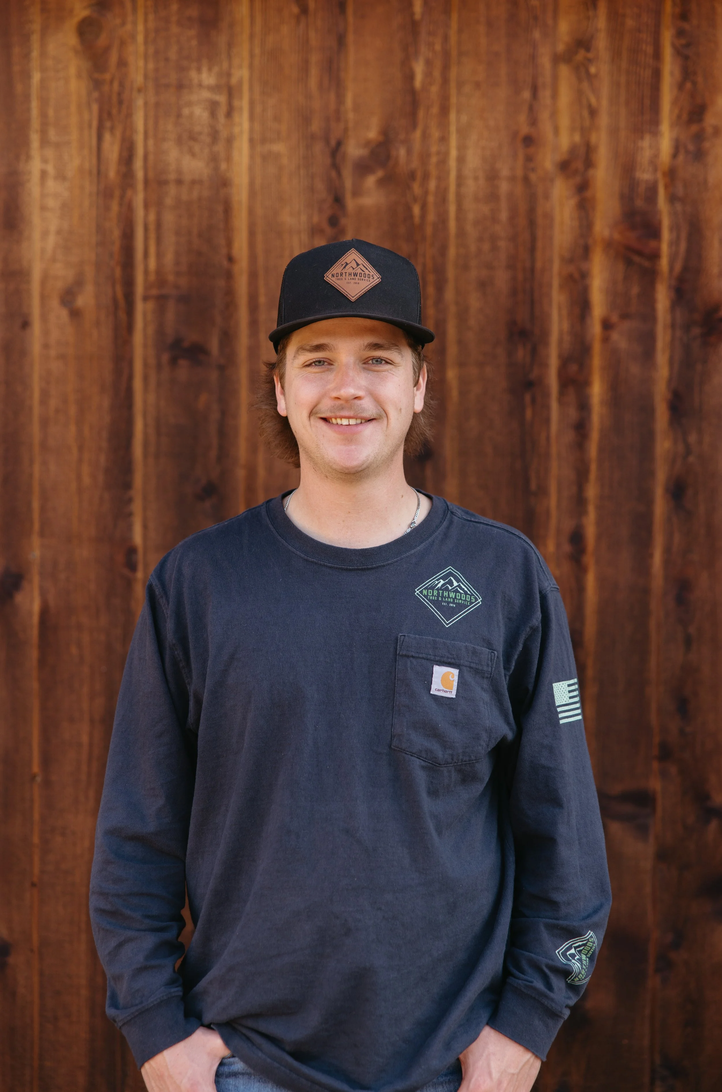 A young man smiling, wearing a black cap and a black long-sleeve shirt with logos, standing in front of a wooden background.