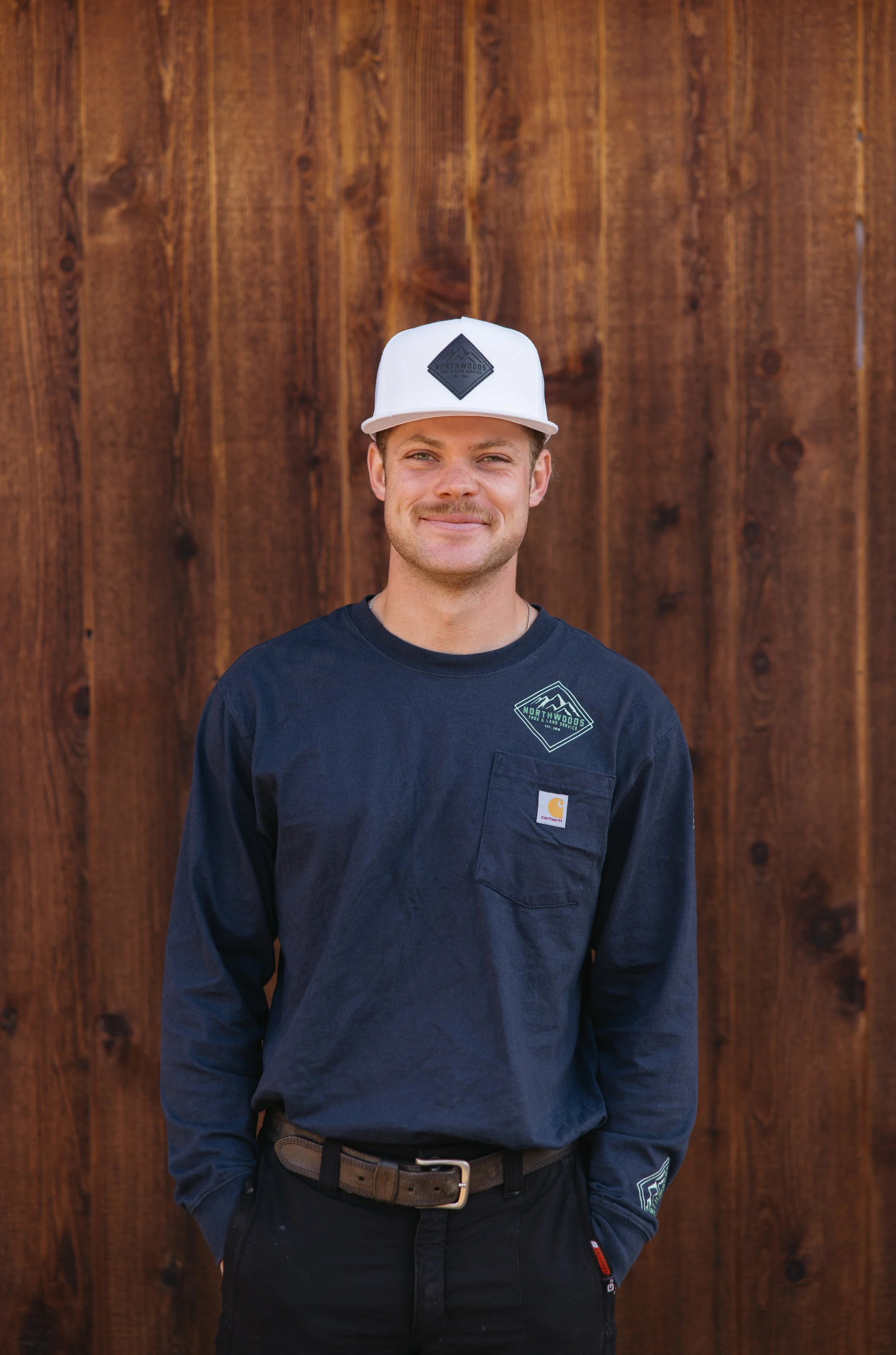 Young man with light brown hair and facial hair, smiling, wearing a white baseball cap, a navy blue Carhartt shirt with a logo on the chest, and standing in front of a wooden background.