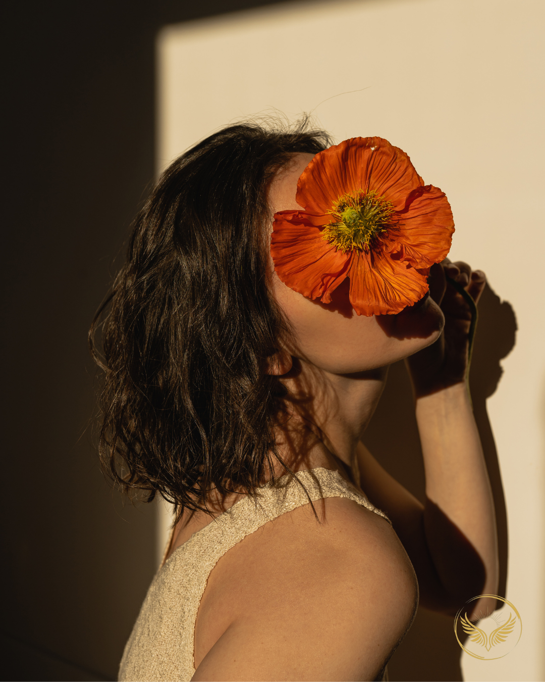 A woman with wavy dark hair holding a large orange flower over her face, casting a shadow on cream-colored background.