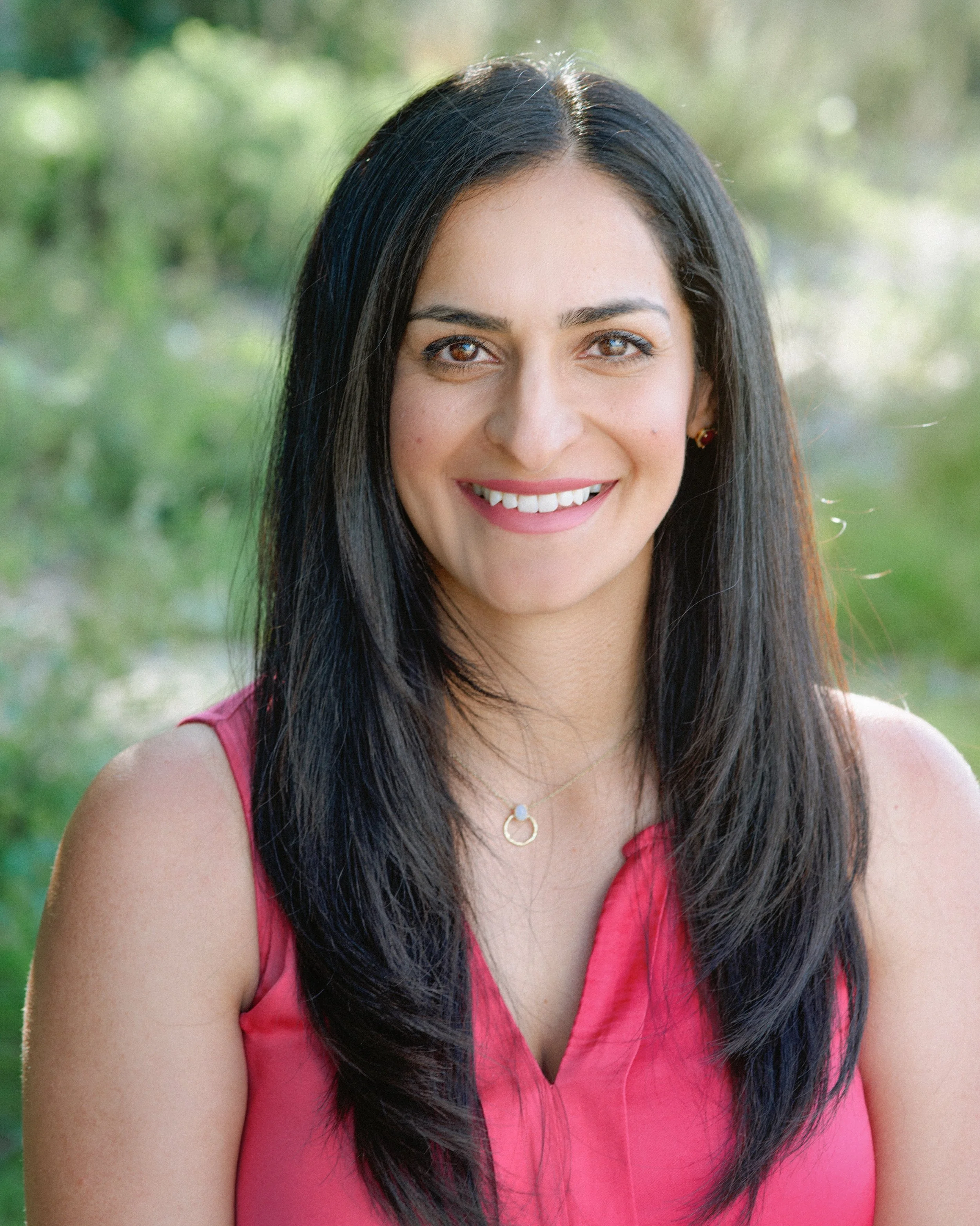Close-up of a woman with long black hair, brown eyes, wearing a pink sleeveless top and a gold necklace, outdoors with blurred greenery background.