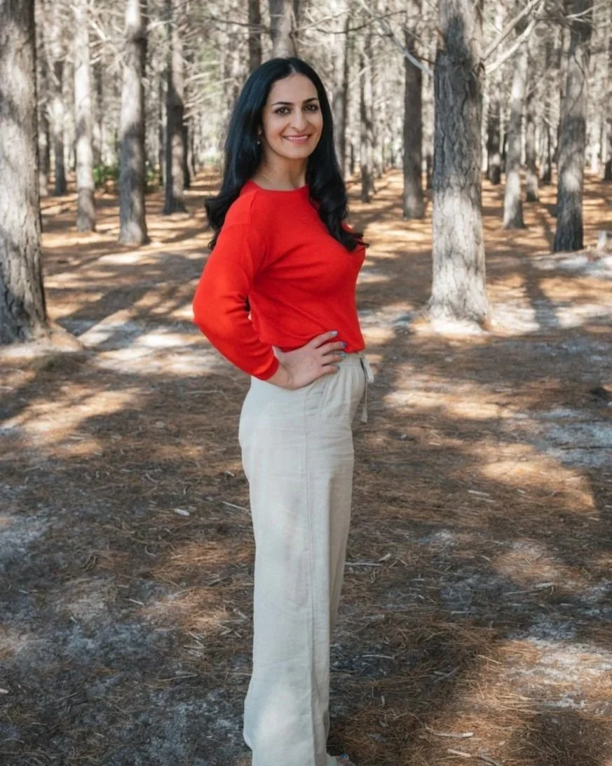 A woman with long black hair and a bright smile posing in a wooded outdoor area with tall trees and patches of sunlight.