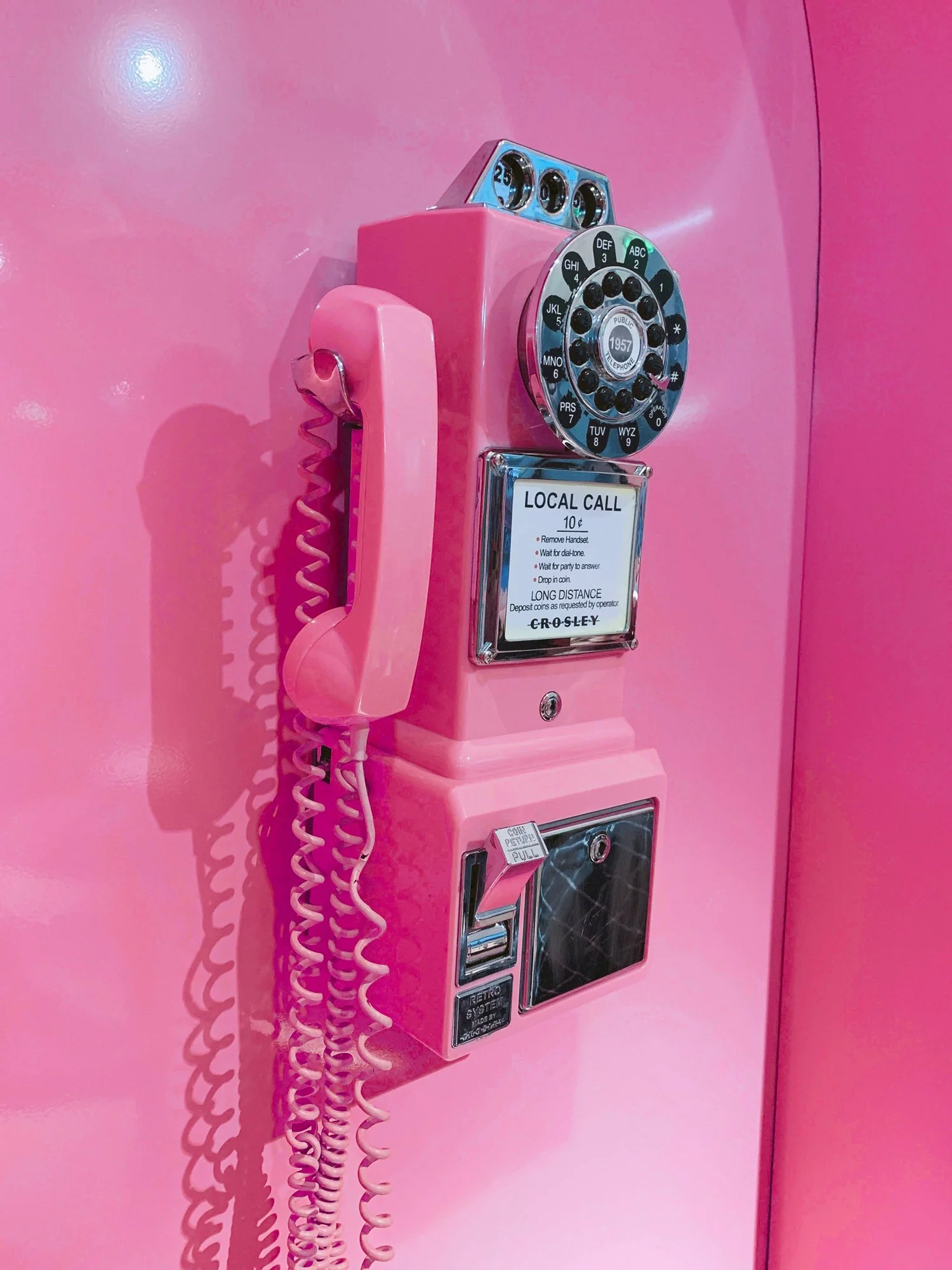 A vintage pink rotary dial phone mounted on a pink wall, with a cord and a coin slot for a payphone, labeled for local calls, typical of mid-20th century design.
