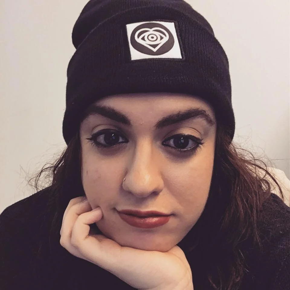A young woman with dark hair wearing a black beanie with a logo, resting her chin on her hand and looking at the camera.