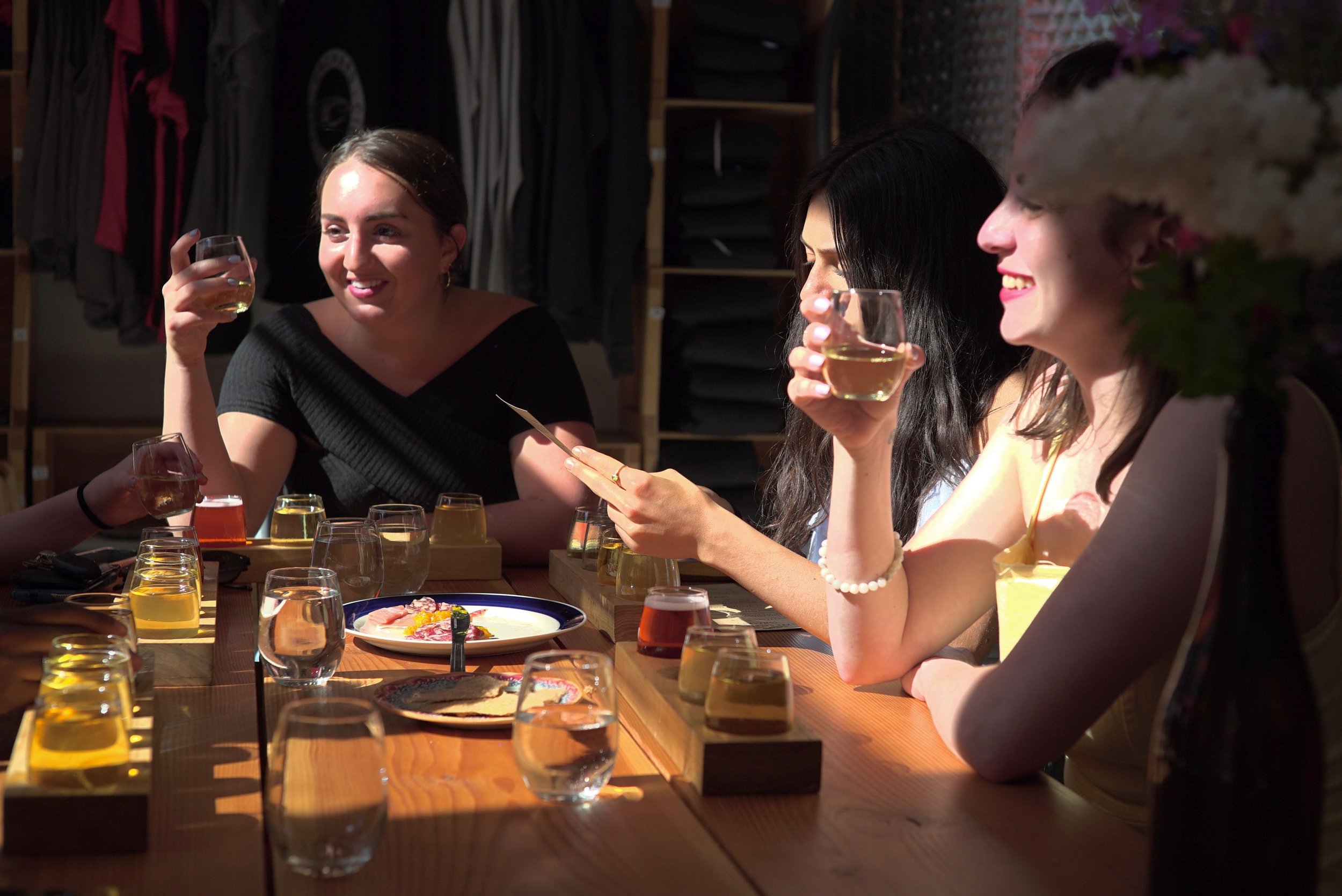 Group of women enjoying cider flight tastings at a wooden table in a sunlit room with glasses and plates.