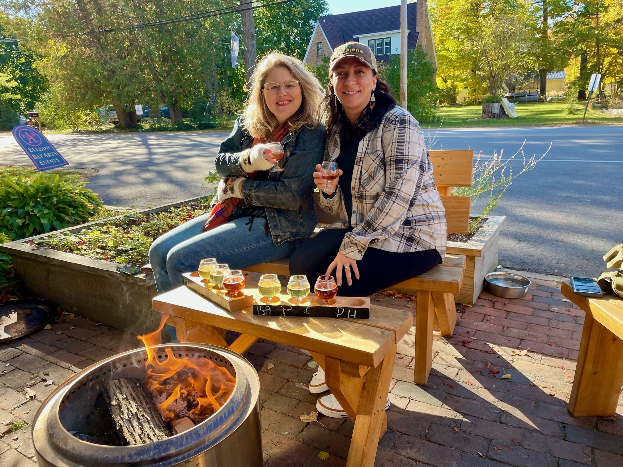 Two people sitting by our outdoor fire pit, enjoying drinks on the patio. They are sitting on wooden benches with a flight of cider in front of them. There is greenery and trees in the background.