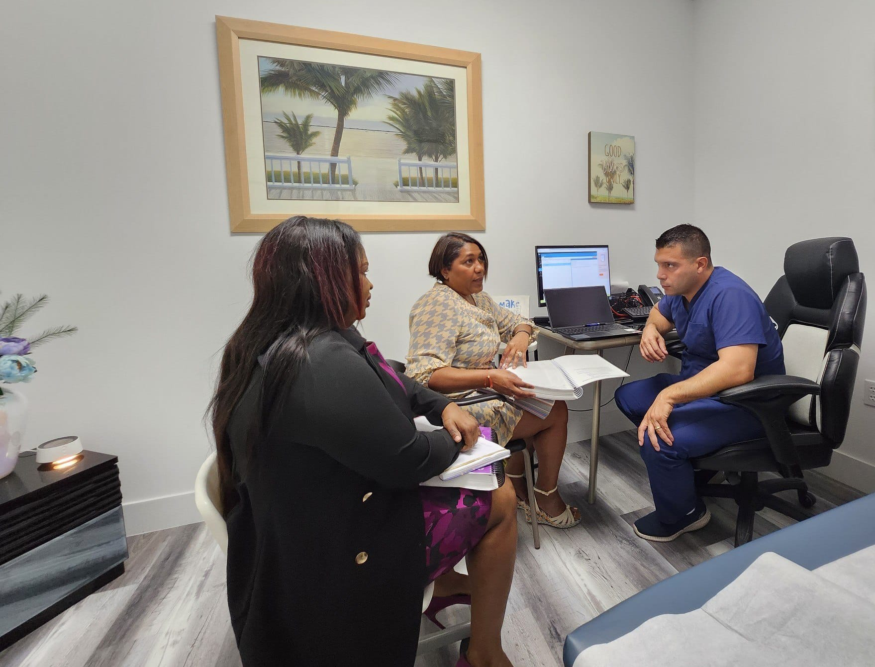 Two women and a man in medical scrubs sitting in a doctor's office, having a conversation. A computer and documents are on the desk.