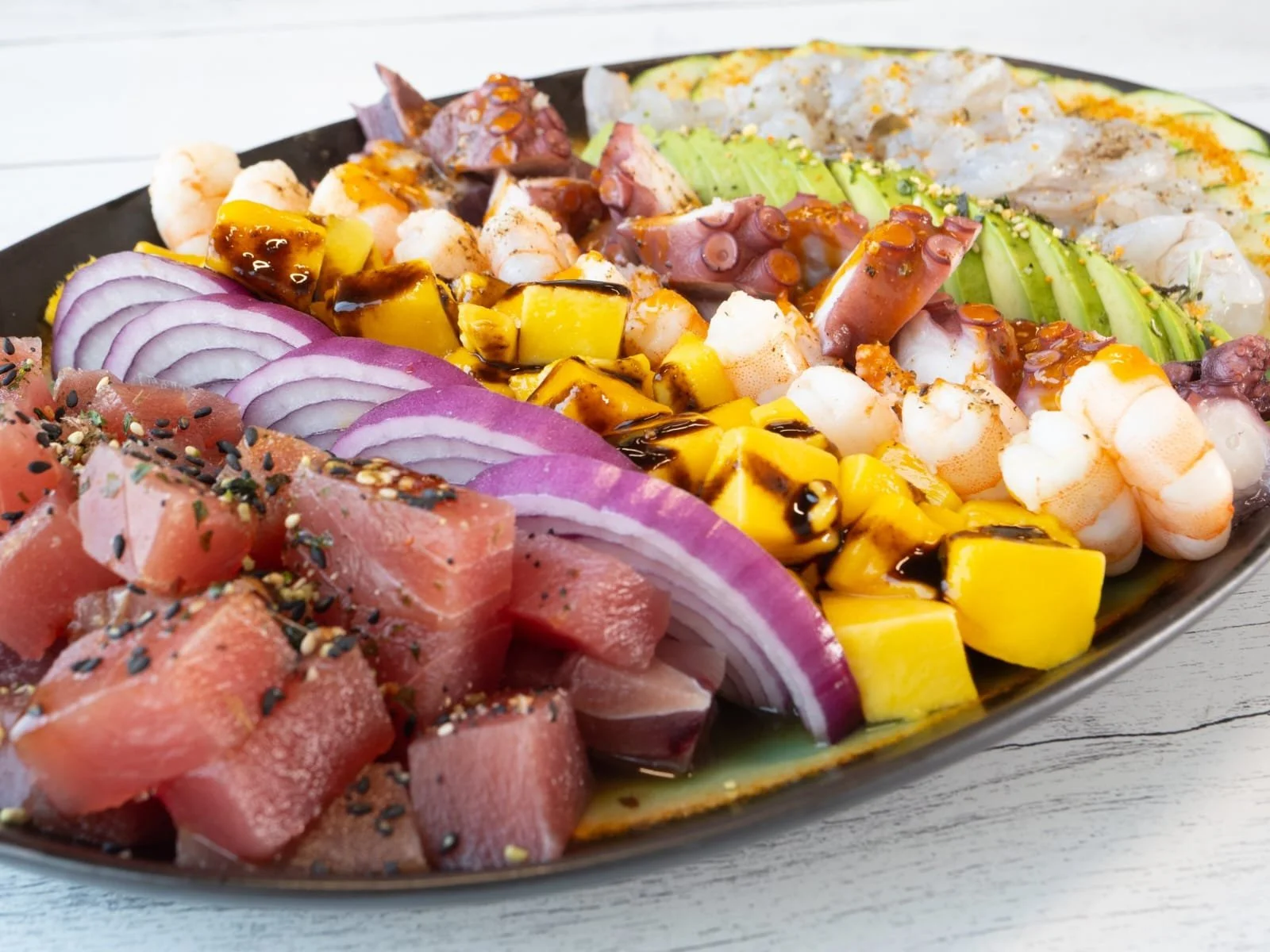Plate of assorted sashimi with tuna, red onion, shrimp, mango, avocado, and drizzle of sauce.
