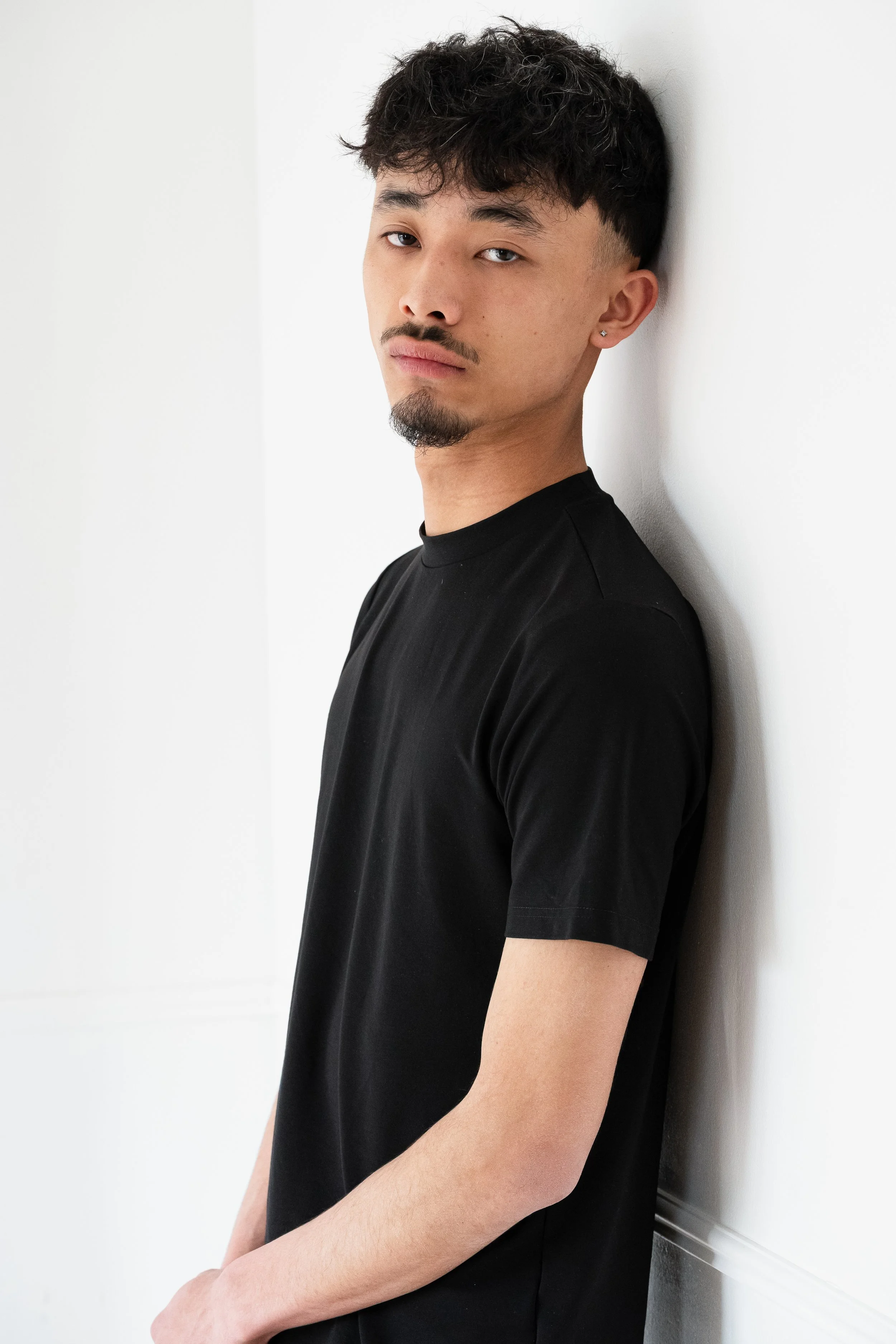 Un homme appuyé contre un mur blanc portant un t-shirt noir.