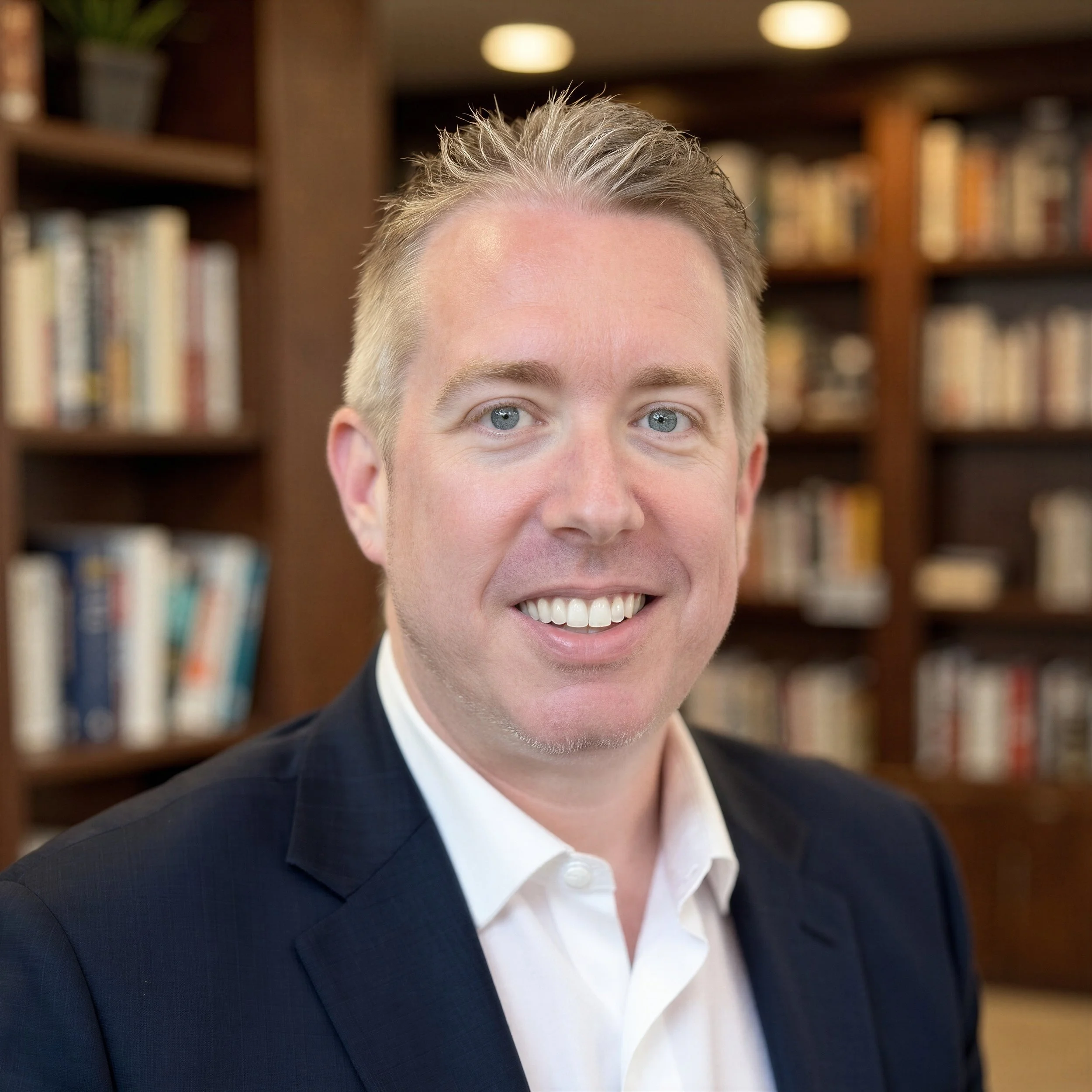 A smiling man with short, styled blonde hair, wearing a dark suit and white shirt, in front of a background of bookshelves.