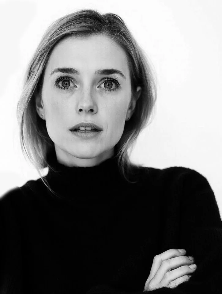A black-and-white portrait of a Clara M. Trenor, Director of Imperfecta Gallery with shoulder-length hair, wearing a black turtleneck sweater, looking directly at the camera with an intense expression, arms crossed.
