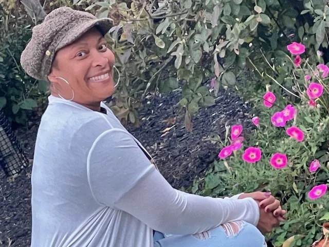 A smiling woman wearing a hat and large hoop earrings, sitting outdoors next to pink flowers and green foliage.