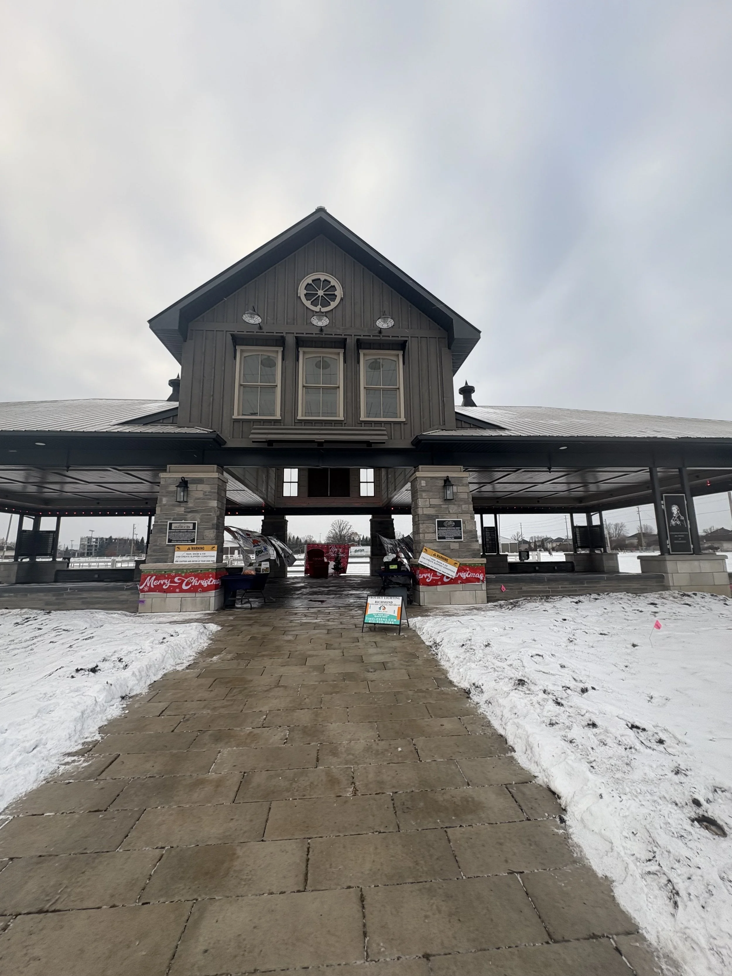 A large gray building with a covered walkway, stone columns, and snow on the ground. Christmas decorations and signs are visible near the entrance.