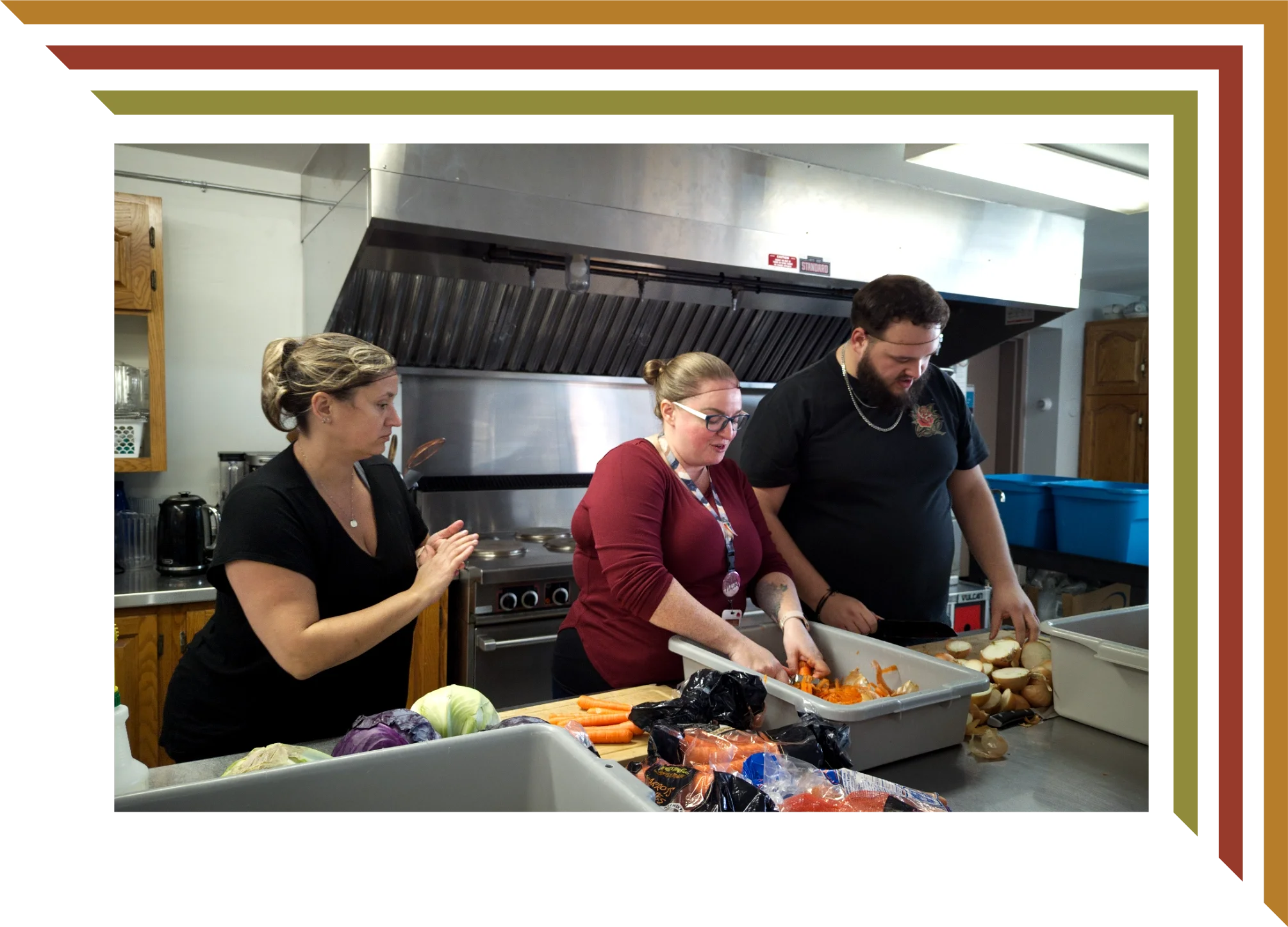 Three people preparing food in a kitchen, chopping vegetables at a stainless steel counter, with a woman on the left clapping her hands, and a woman and man on the right focused on their tasks.