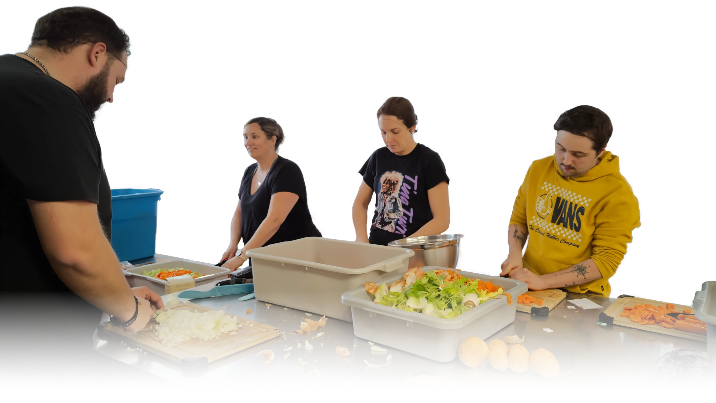 Four people preparing vegetables in a kitchen or community cooking space. They are chopping and organizing vegetables like onions, carrots, and leafy greens on a stainless steel table.