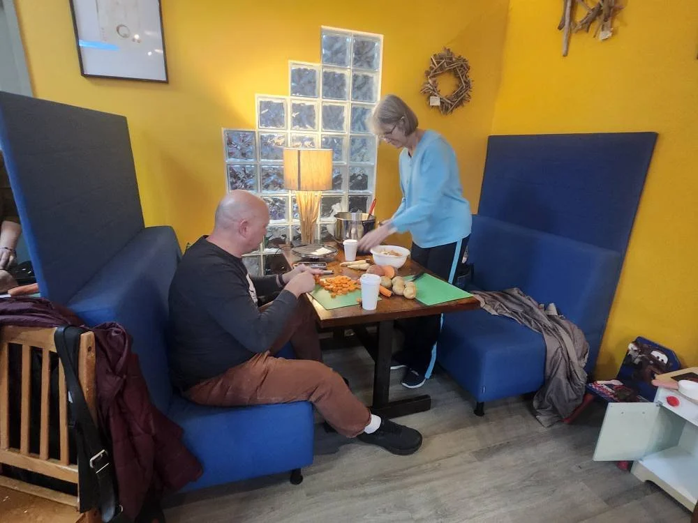 A man and woman are in a cozy restaurant, preparing and serving a meal at a table with a green cutting board, vegetables, and disposable cups. The man is seated, while the woman is standing, wearing a light blue sweater. The restaurant has yellow walls, blue booth seating, and decorative flower wreaths.