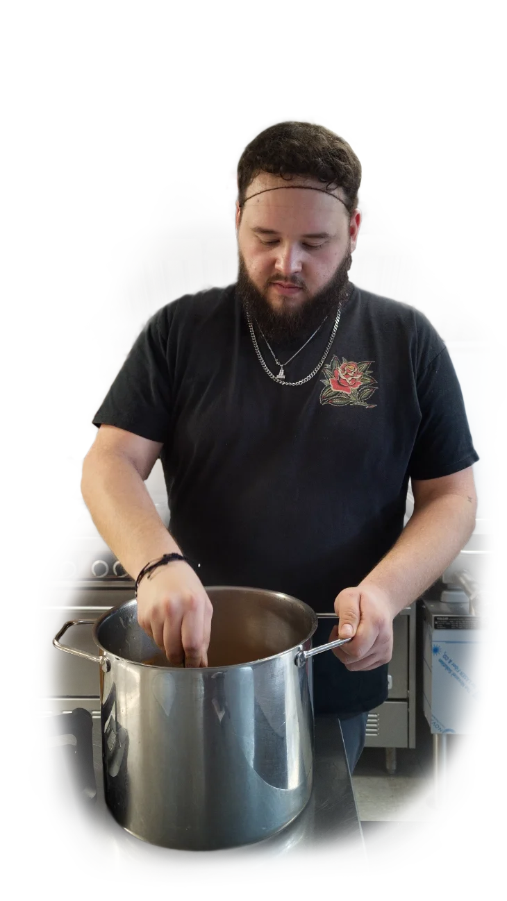 A man with a beard and dark hair wearing a black T-shirt with a rose embroidery, a chain necklace, and a headband, stirring a large stainless steel pot on a stove in a commercial kitchen.