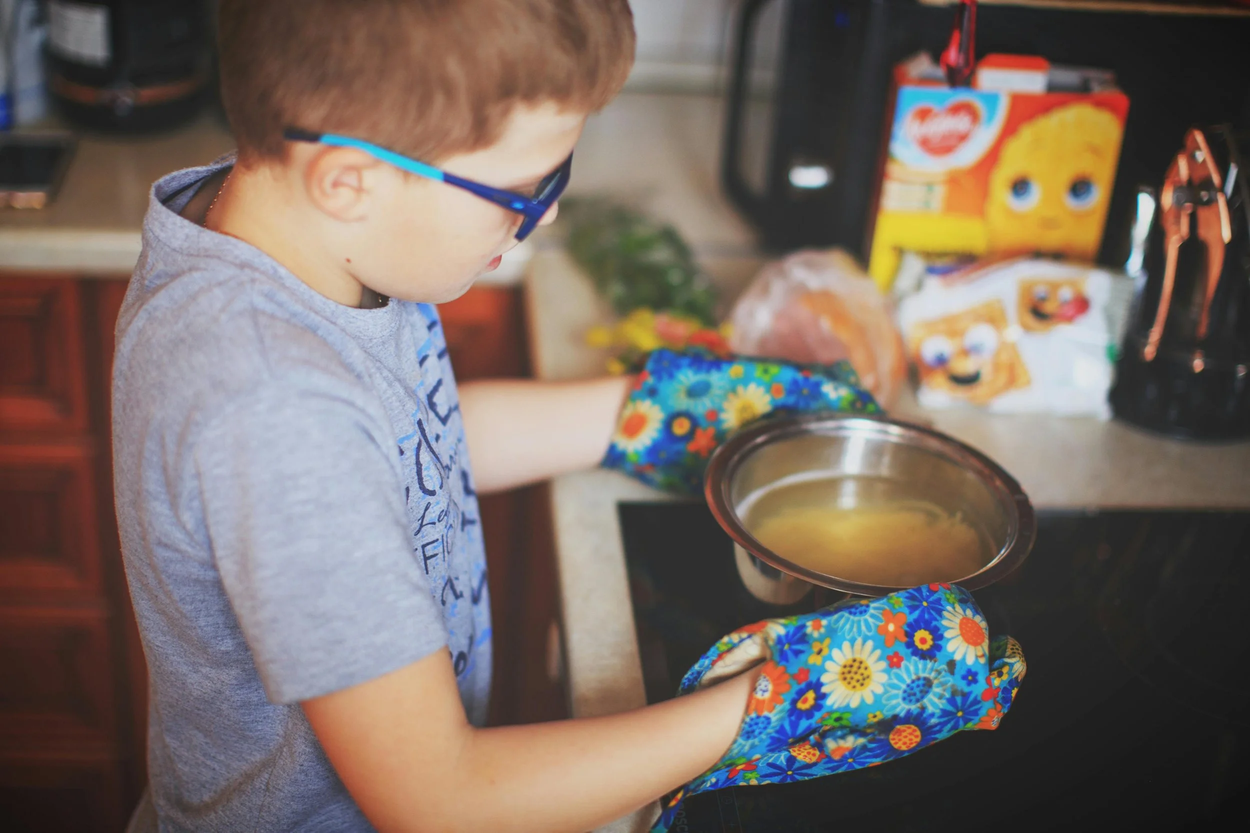 A young boy wearing glasses and a gray t-shirt, with floral oven mitts, is cooking on a stove, holding a metal pot with a yellow liquid inside.