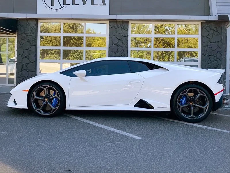 A white luxury sports car, a Lamborghini Huracan, parked in a parking lot in front of a building with large windows and stone walls.