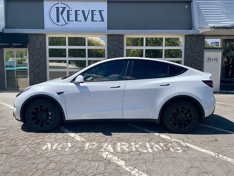 A white Tesla Model Y parked in a marked parking space labeled 'NO PARKING' in front of a building with the sign 'KEEVES'.