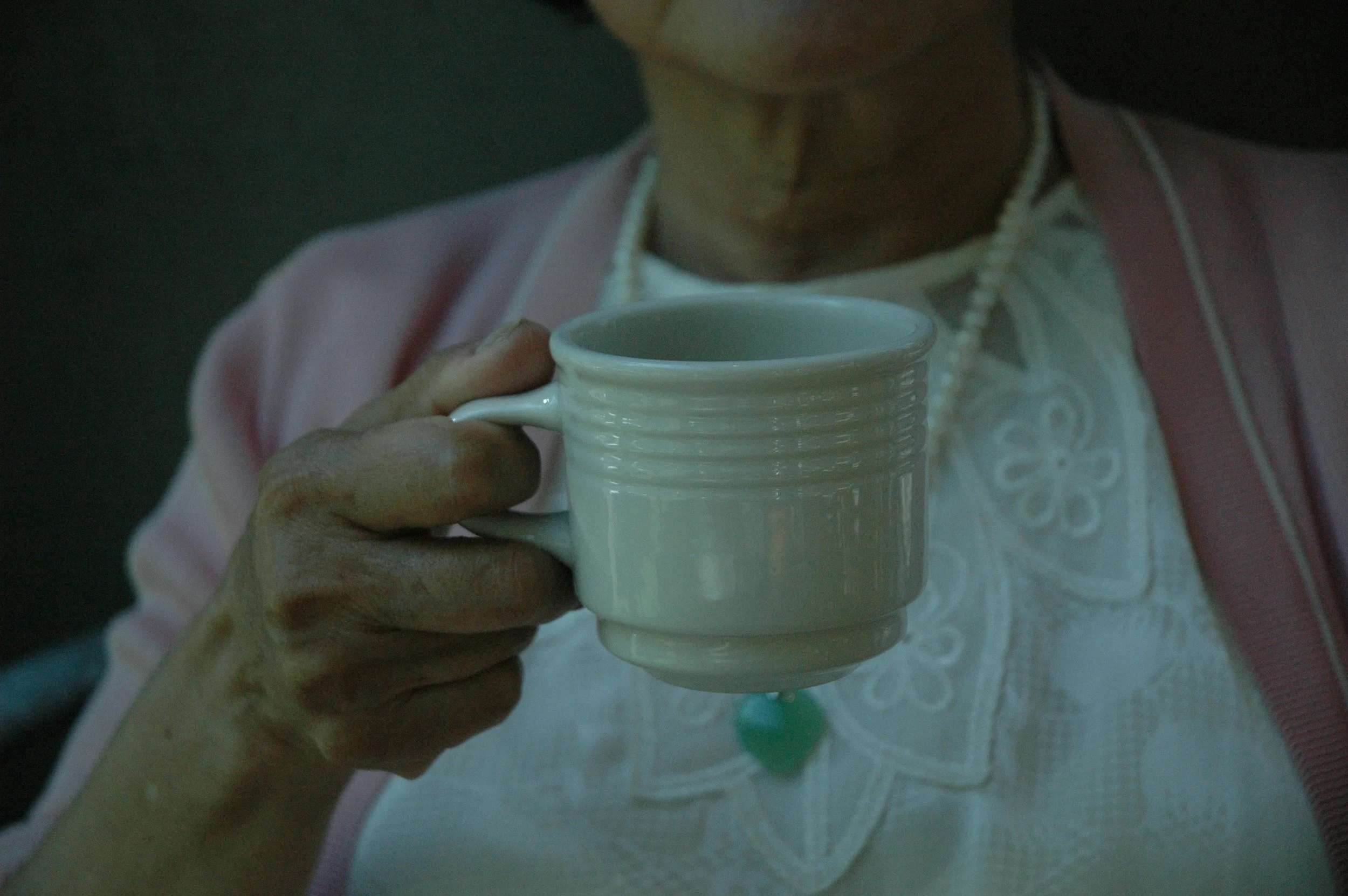 "Older woman wearing a pink cardigan and pearls, gently holding a white mug—representing hydration support with dignity for those with dementia."