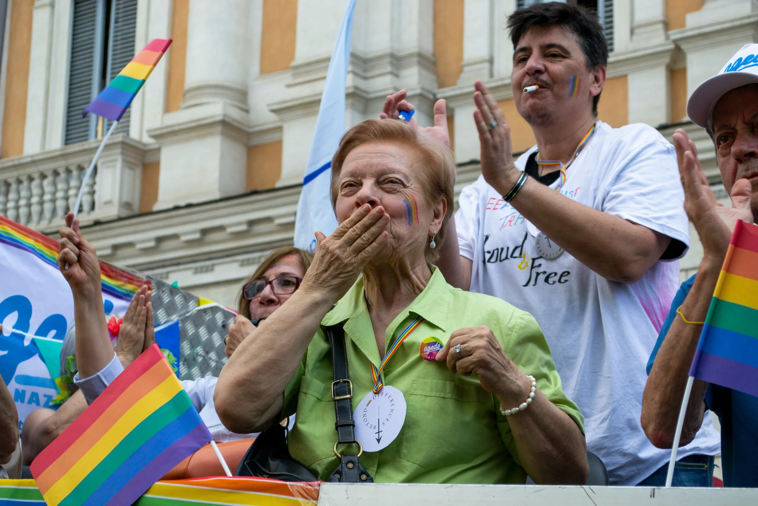 An older adult wearing a rainbow lanyard and green shirt blows a kiss at a Pride celebration, surrounded by others holding rainbow flags and cheering.