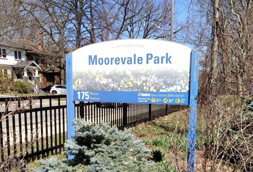 Sign for Moorevale Park with blue text on white background, trees, houses, and parked cars in the background.