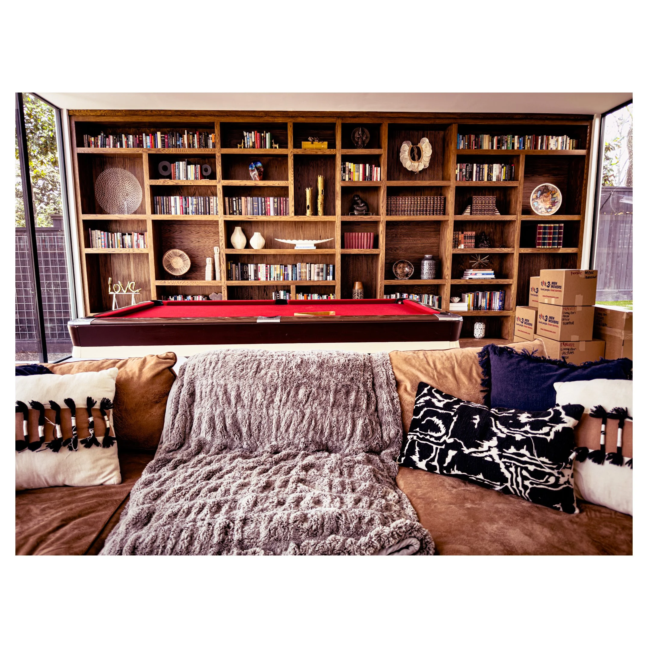 Living room with brown sofa, decorative pillows, fuzzy throw blanket, and a red pool table in front of a large wooden bookshelf filled with books and decorative items.