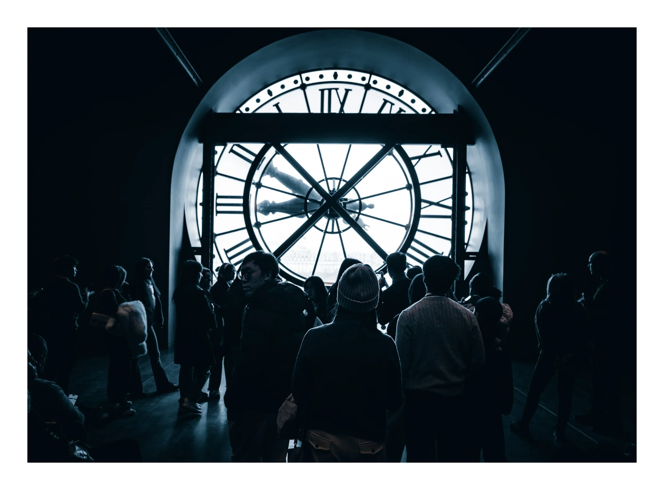 People gathered inside a building near a large clock face with Roman numerals, viewed from the inside, with silhouettes of individuals against a backlit window.