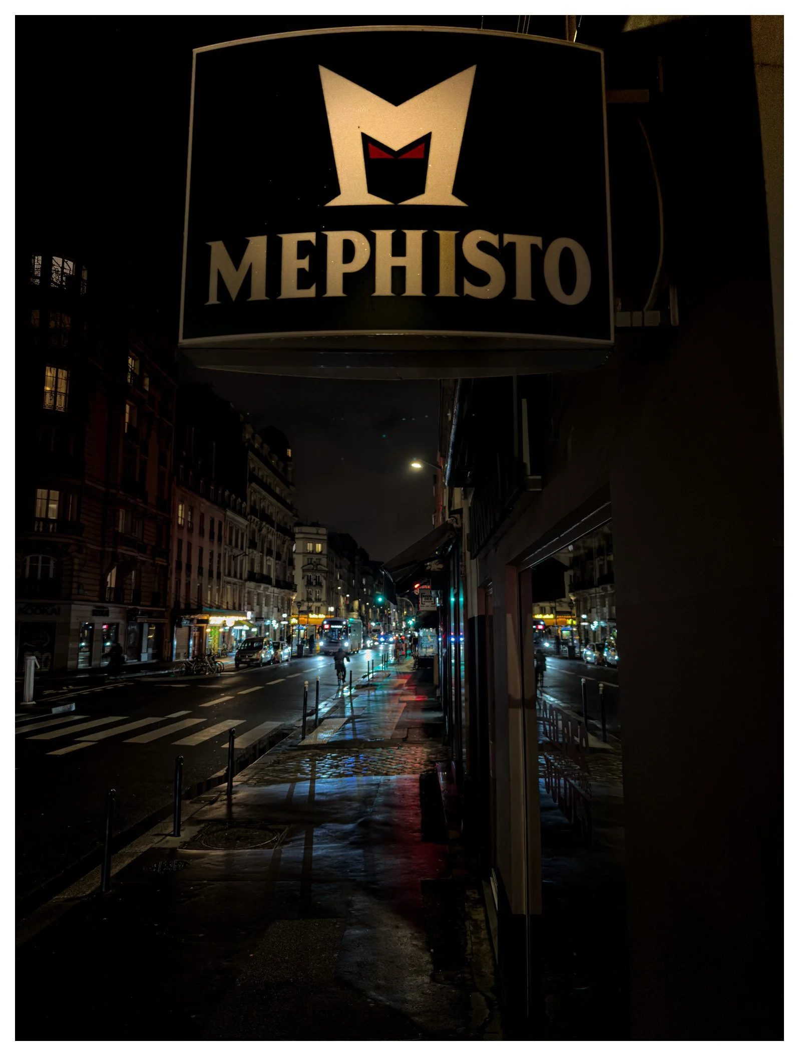 Nighttime city street scene with illuminated shop signs, parked cars, and a bus, seen from the sidewalk in front of a store with a sign that reads 'Mephisto'.