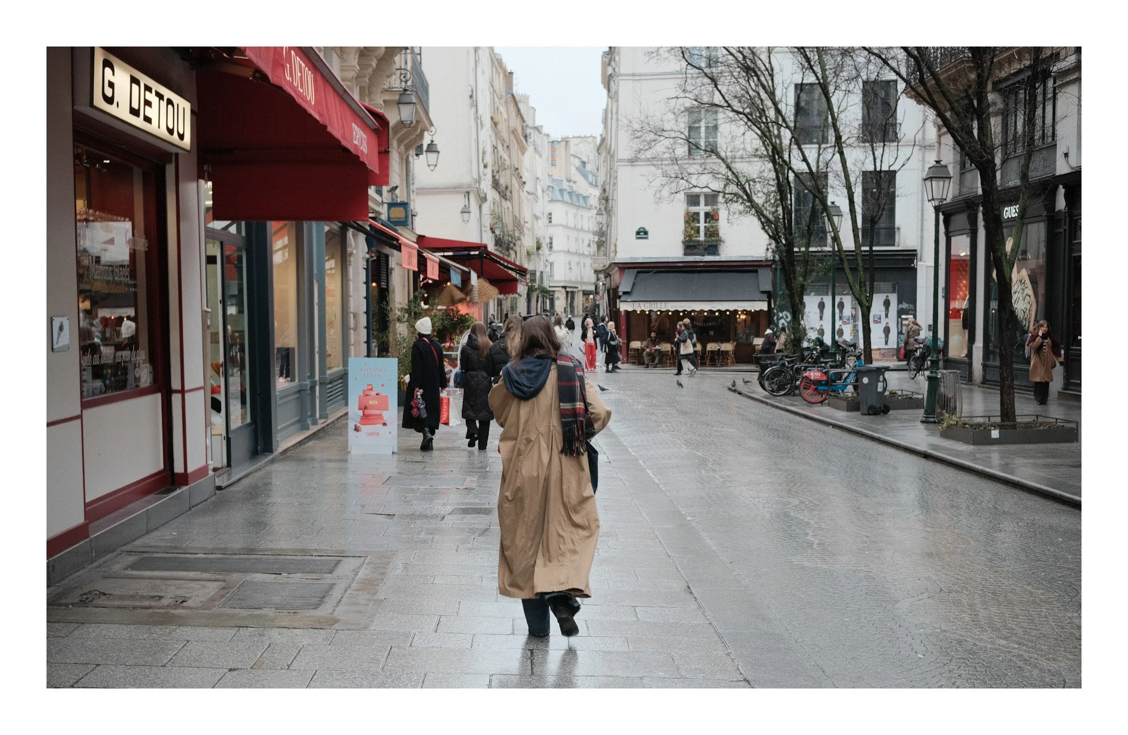 Street scene in Paris with pedestrians on a wet sidewalk, storefronts, trees, and outdoor seating in the background.