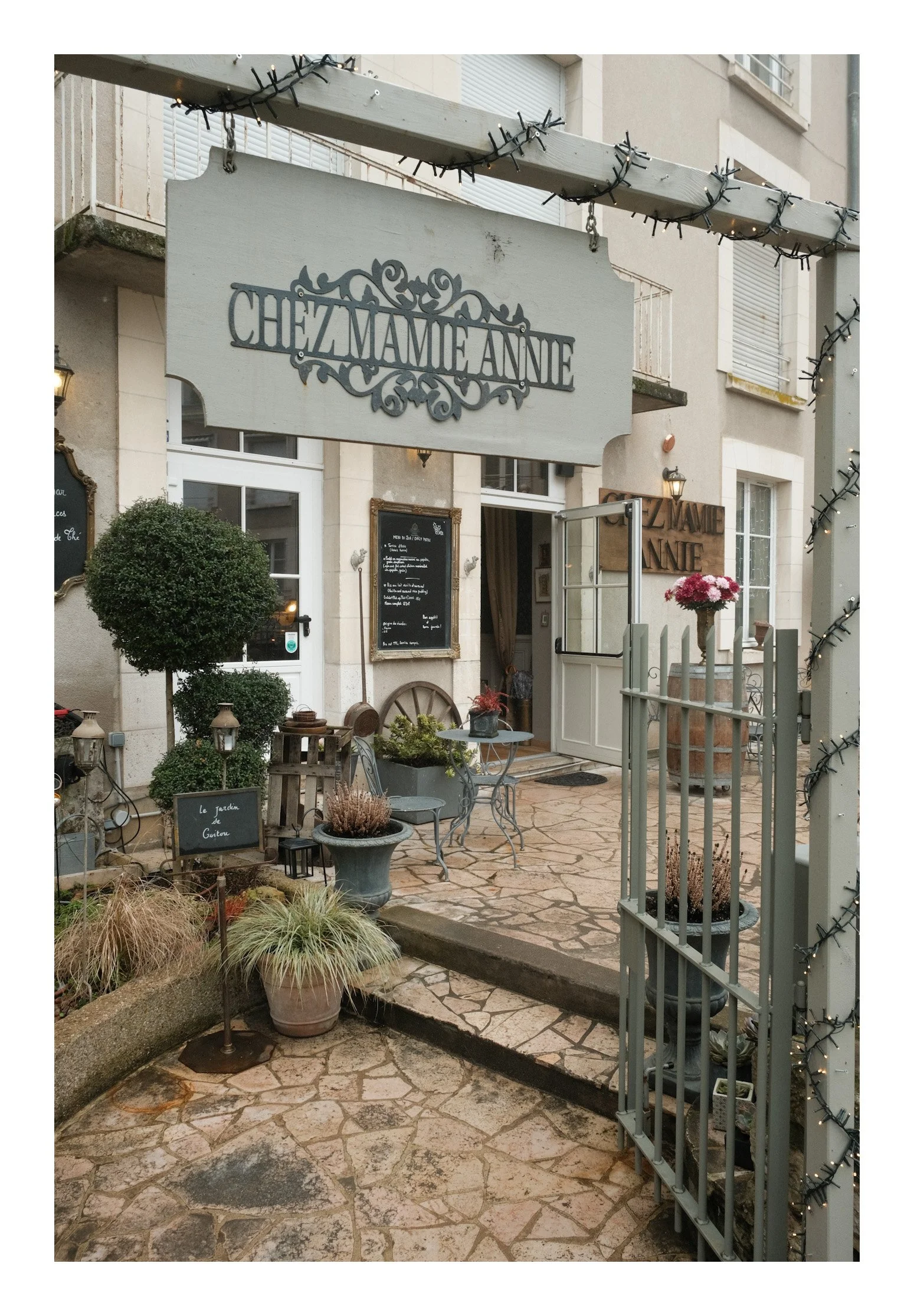 Exterior view of a European-style restaurant or café with signs reading "Chez Mamie Annie" and outdoor seating, potted plants, and garden décor.