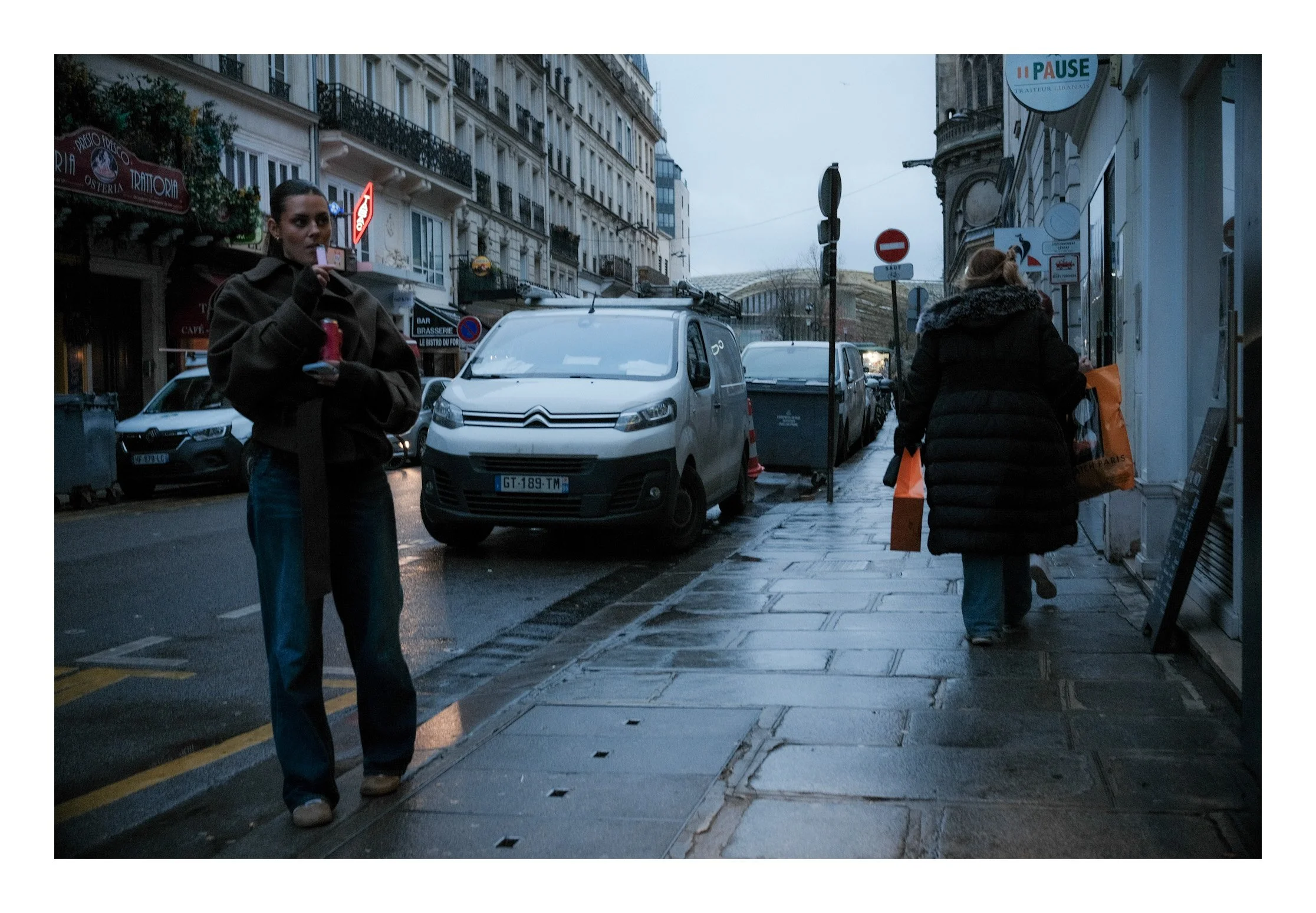 A rainy street scene in a city, with two women walking on the wet sidewalk. One woman is on the left, holding a pink object and a red cup, wearing a dark jacket and jeans. The other woman is on the right, carrying orange shopping bags, dressed in a b