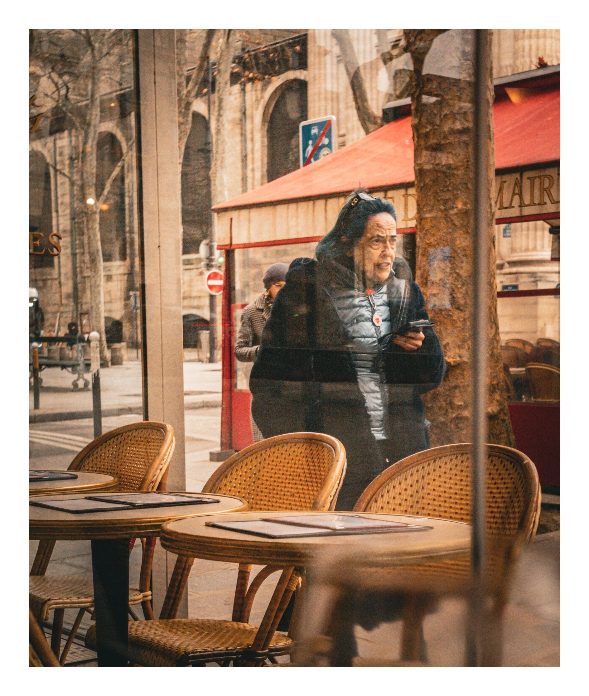 View through a restaurant window showing a woman outside looking at her phone, with outdoor tables and chairs in the foreground, and a street scene with trees, a red storefront awning, and pedestrians in the background.