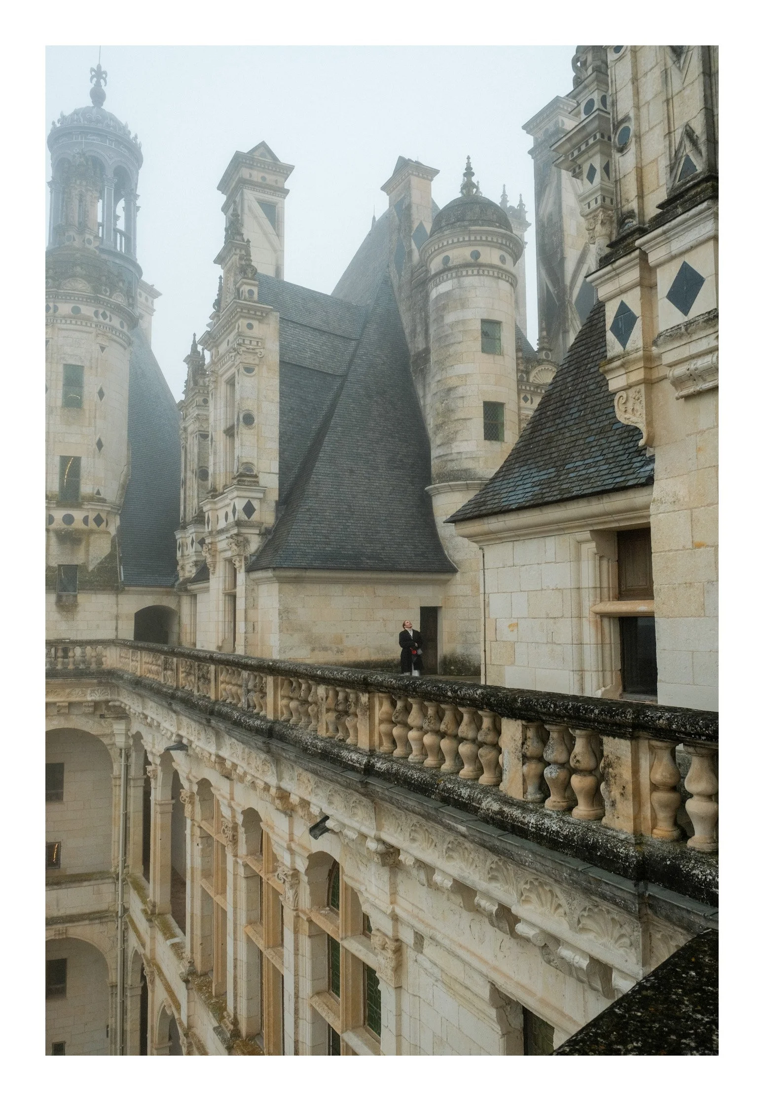 A historic castle with turrets and ornate architecture, shrouded in fog, with a person standing on a balcony.
