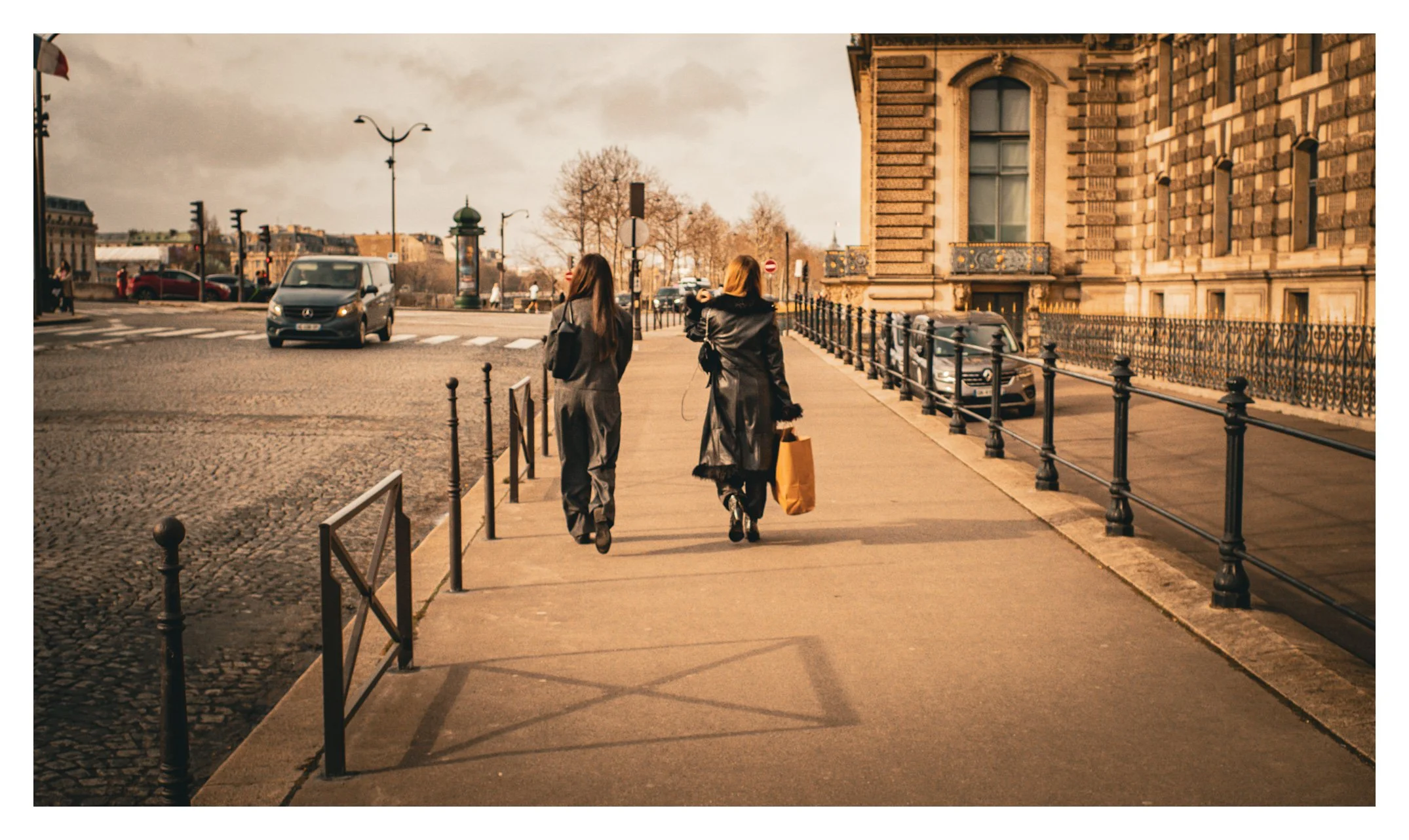 Two women walking on a city sidewalk past old brick buildings, with parked cars and a street crossing ahead under a cloudy sky during daytime.