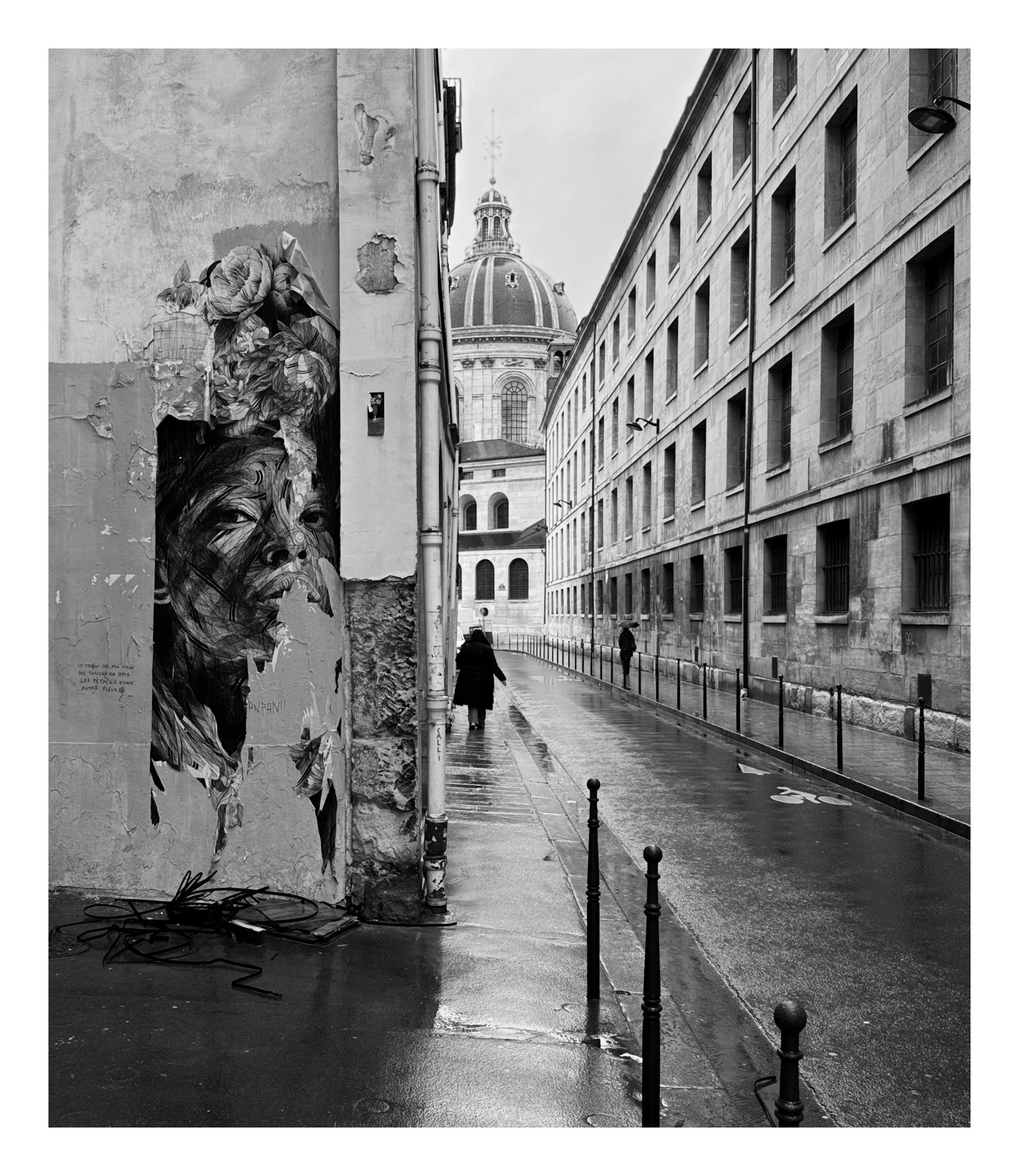 Black and white photo of a wet city street with two pedestrians walking and a large dome building in the background. A mural of a woman's face with flowers is on a wall on the left side.
