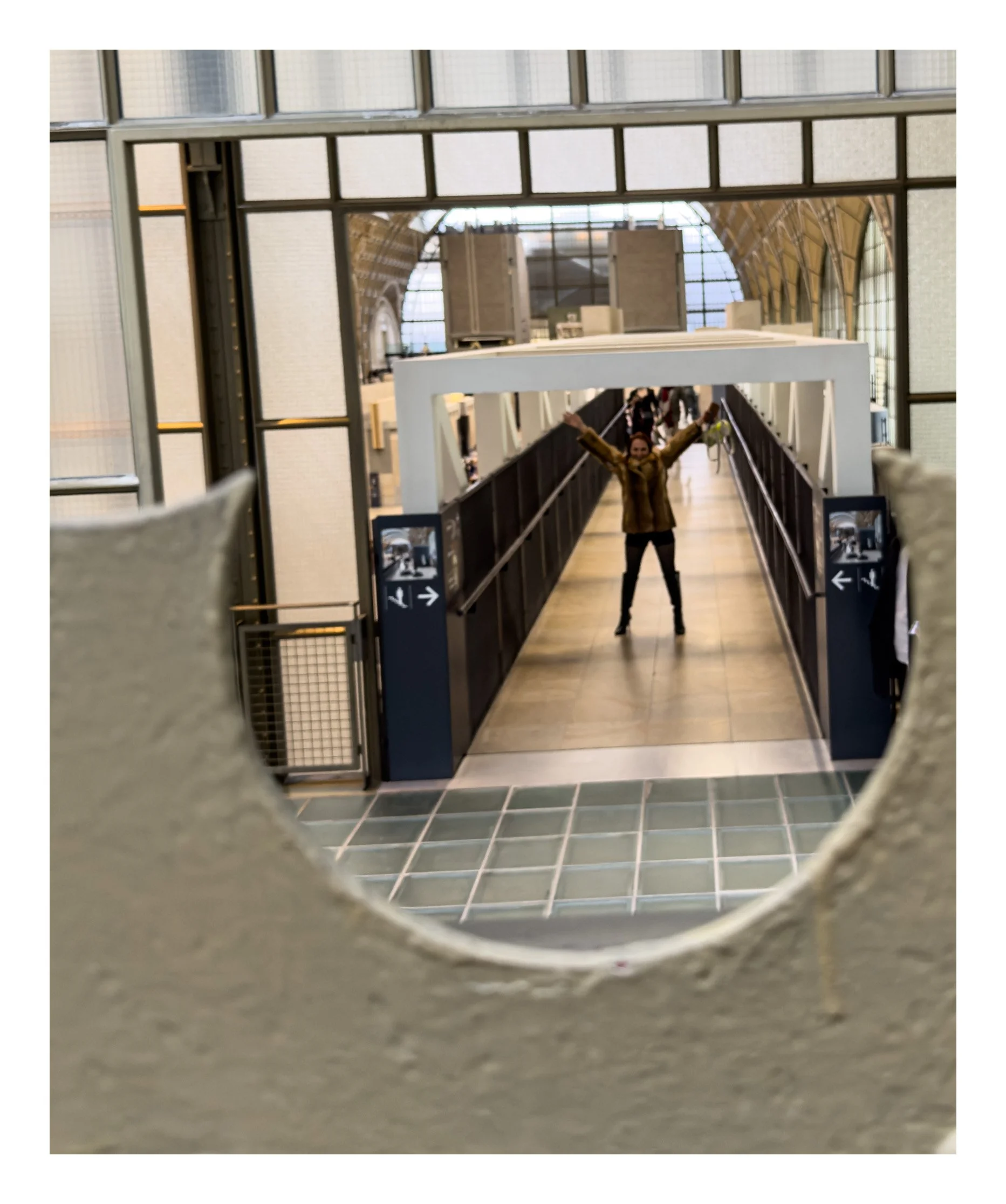 A person is taking a photo in a mirror at an airport, with their arms raised in a 'V' shape, as seen through a circular hole in the foreground wall.